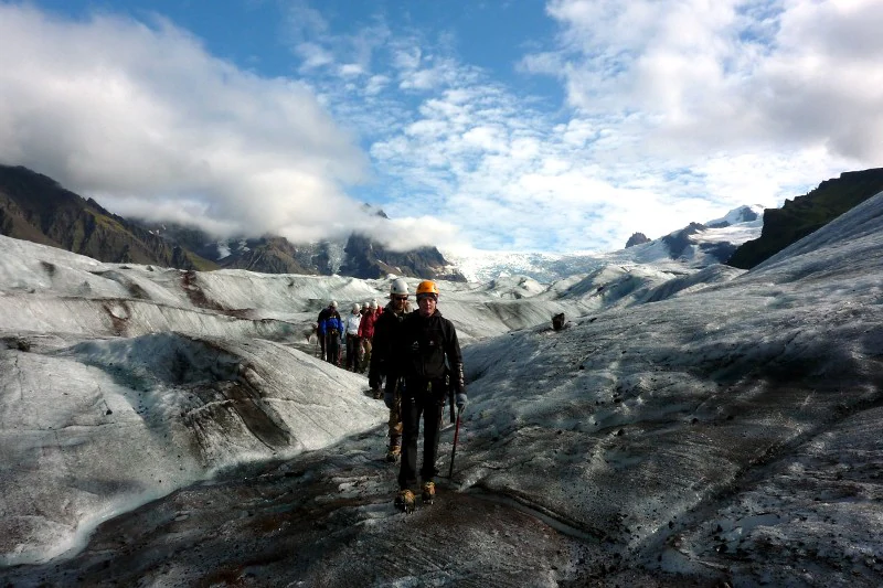 A group of hikers walking on a glacier with mountains in the background under a partly cloudy sky