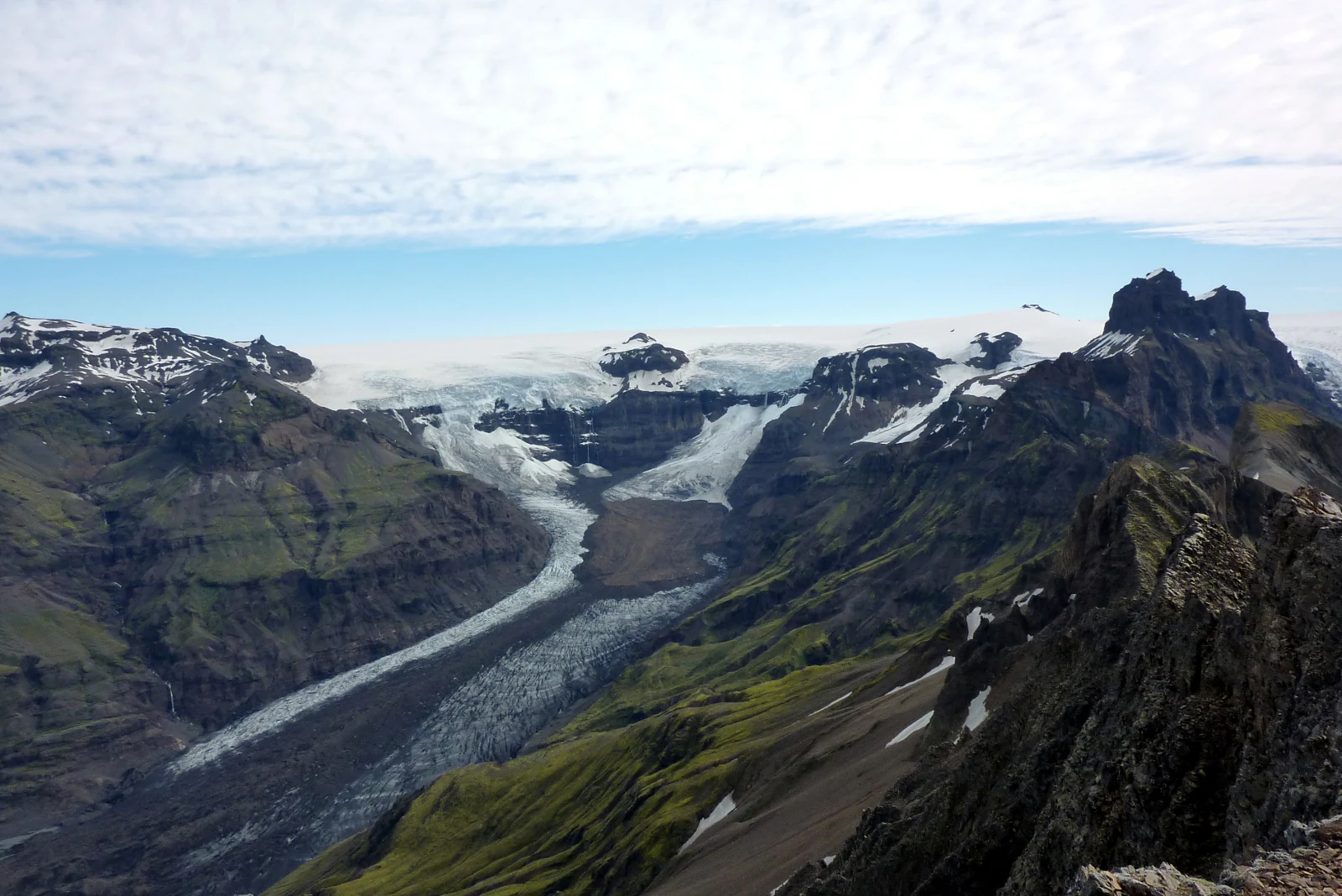 Panoramic view of a glacier flowing through a mountain valley with rocky peaks, grassy slopes, and snow patches under partly cloudy sky