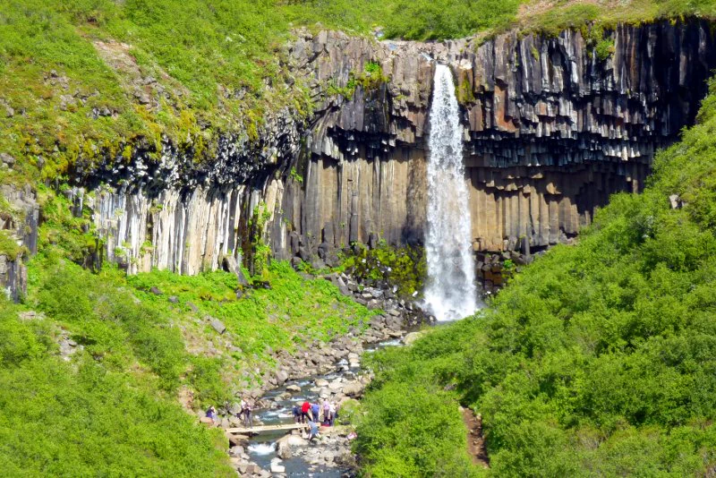 A waterfall cascading down a rocky cliff surrounded by lush green vegetation with a small group of people standing near the stream below