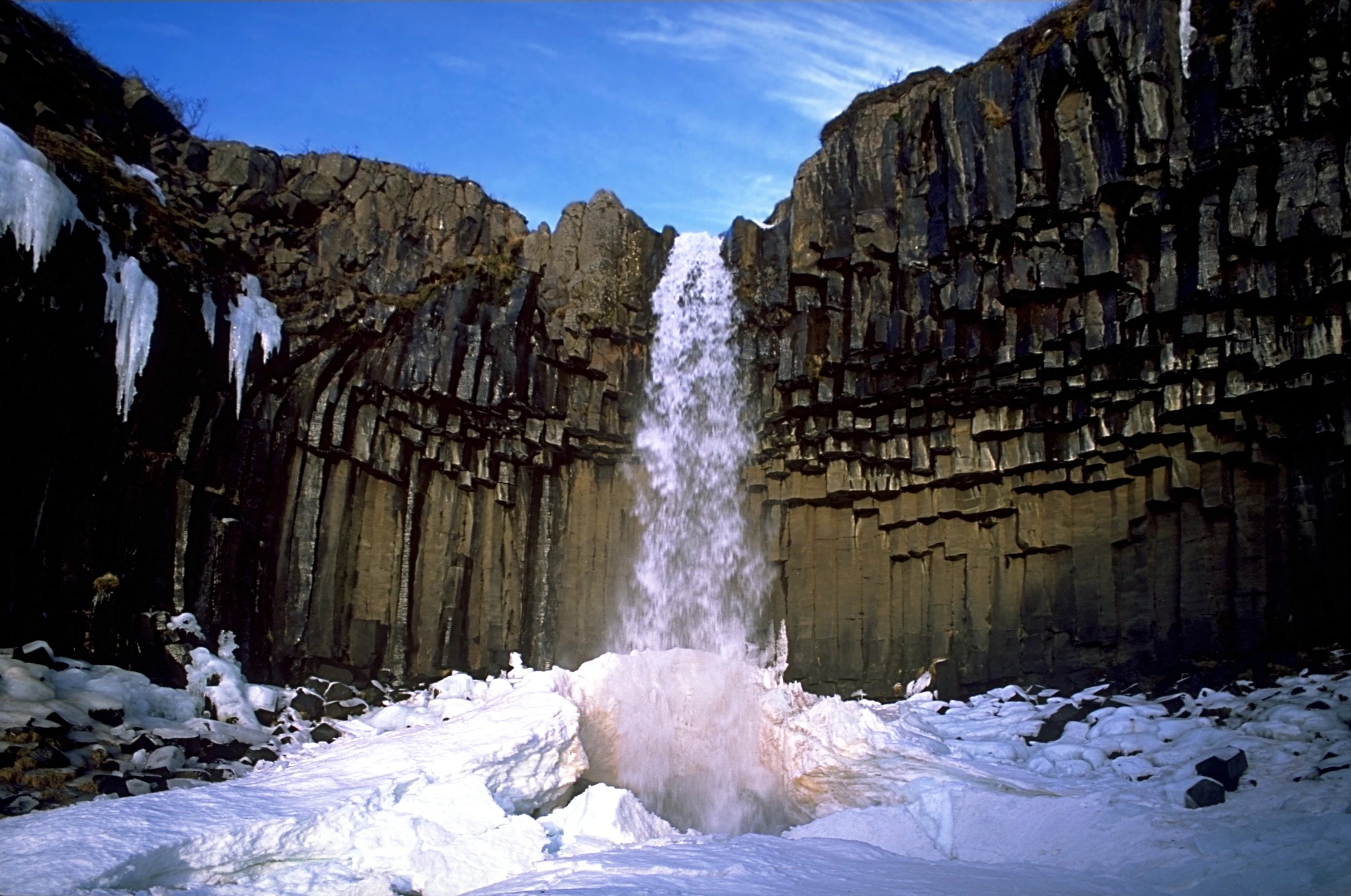 Waterfall cascading between basalt columns with snow-covered ground and ice formations under a clear blue sky