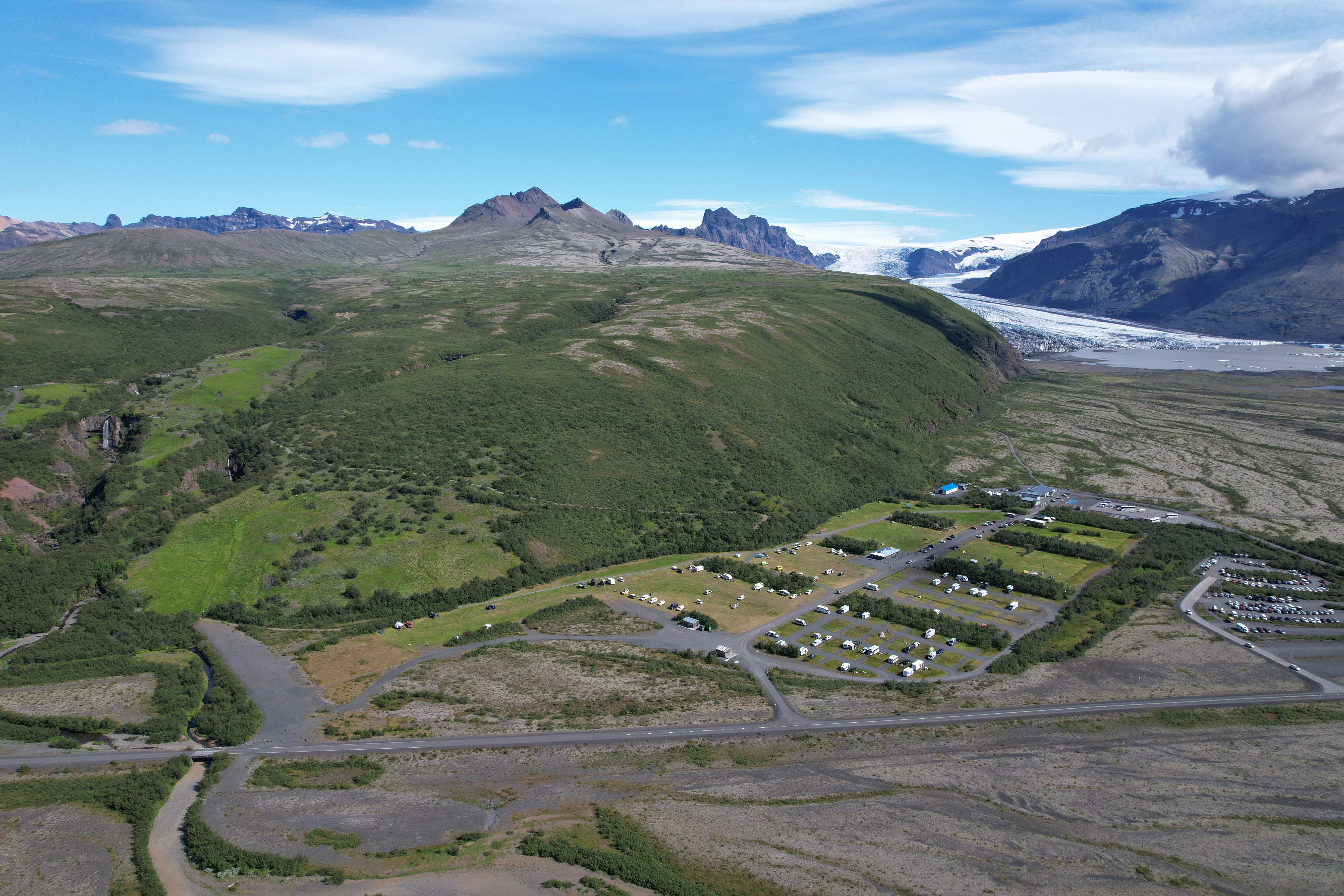 Aerial view of Skaftafell National Park showing a campsite with tents, roads, a glacier, and waterfall under a partly cloudy sky