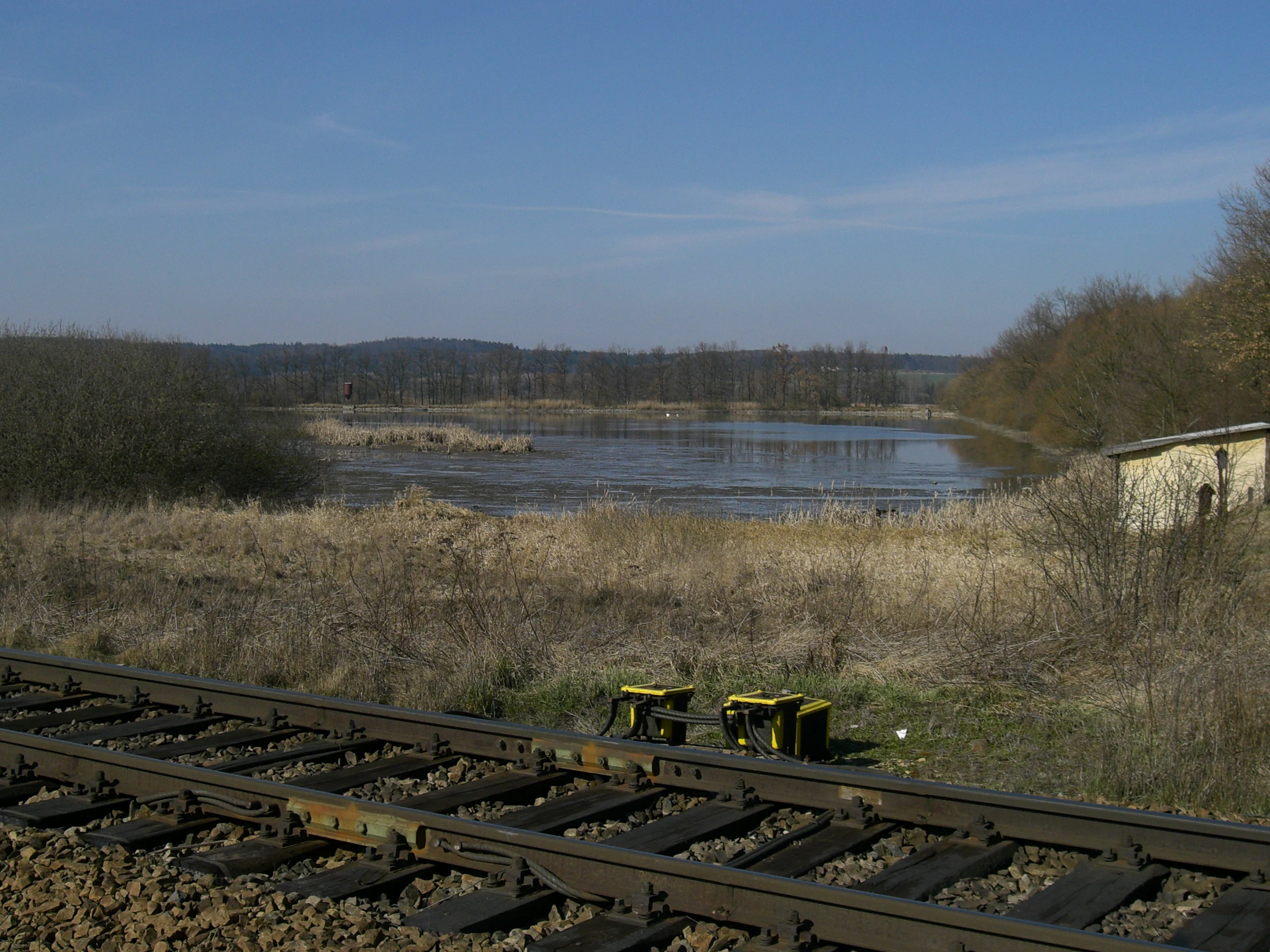 A view of a pond with calm water, dry marshy vegetation in the foreground, a small rustic building on the right, and railroad tracks in the immediate foreground.
