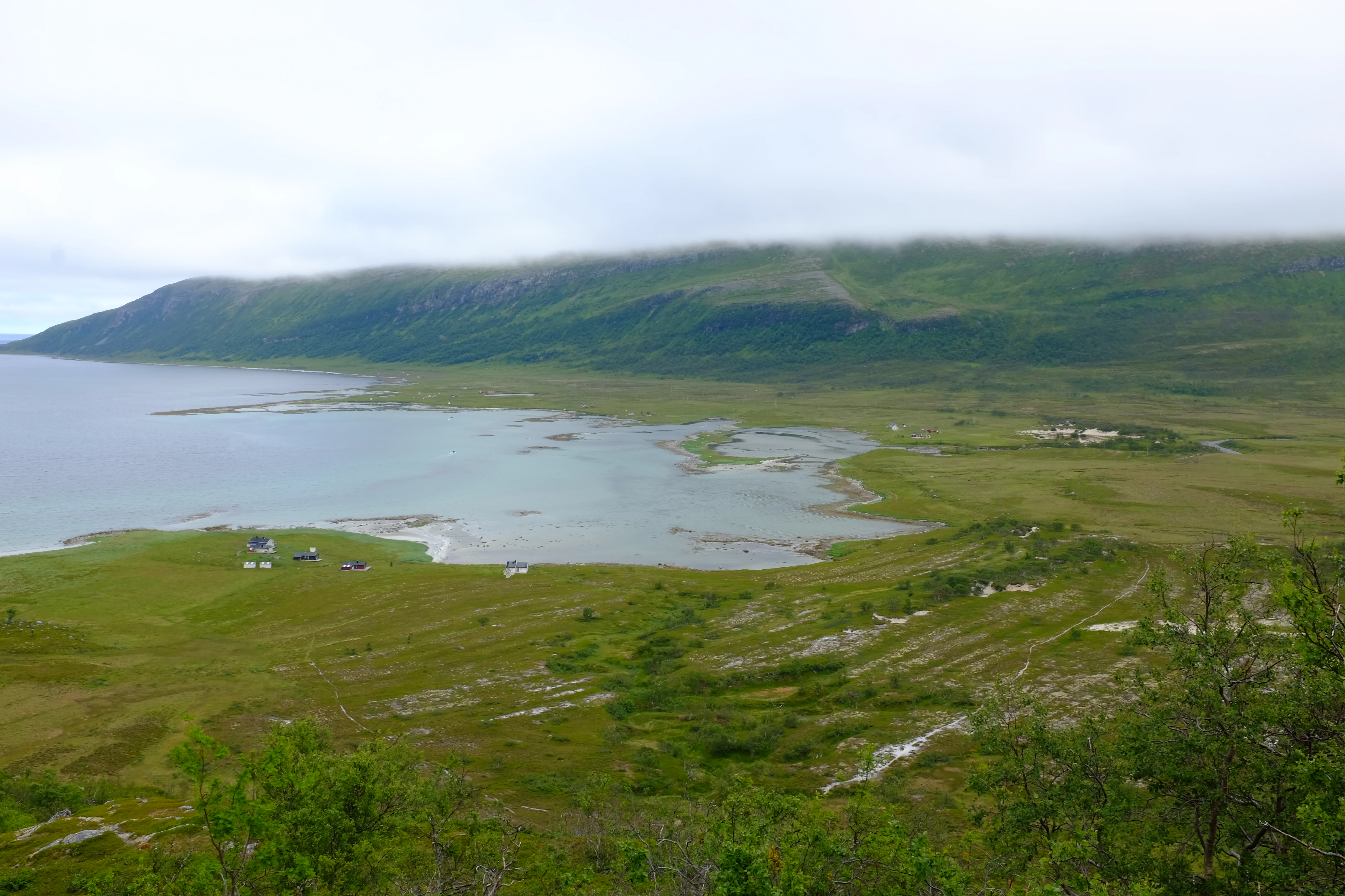 A high-angle view of a calm bay with grassy shores, small buildings in the distance, and low-lying mountains covered in clouds.