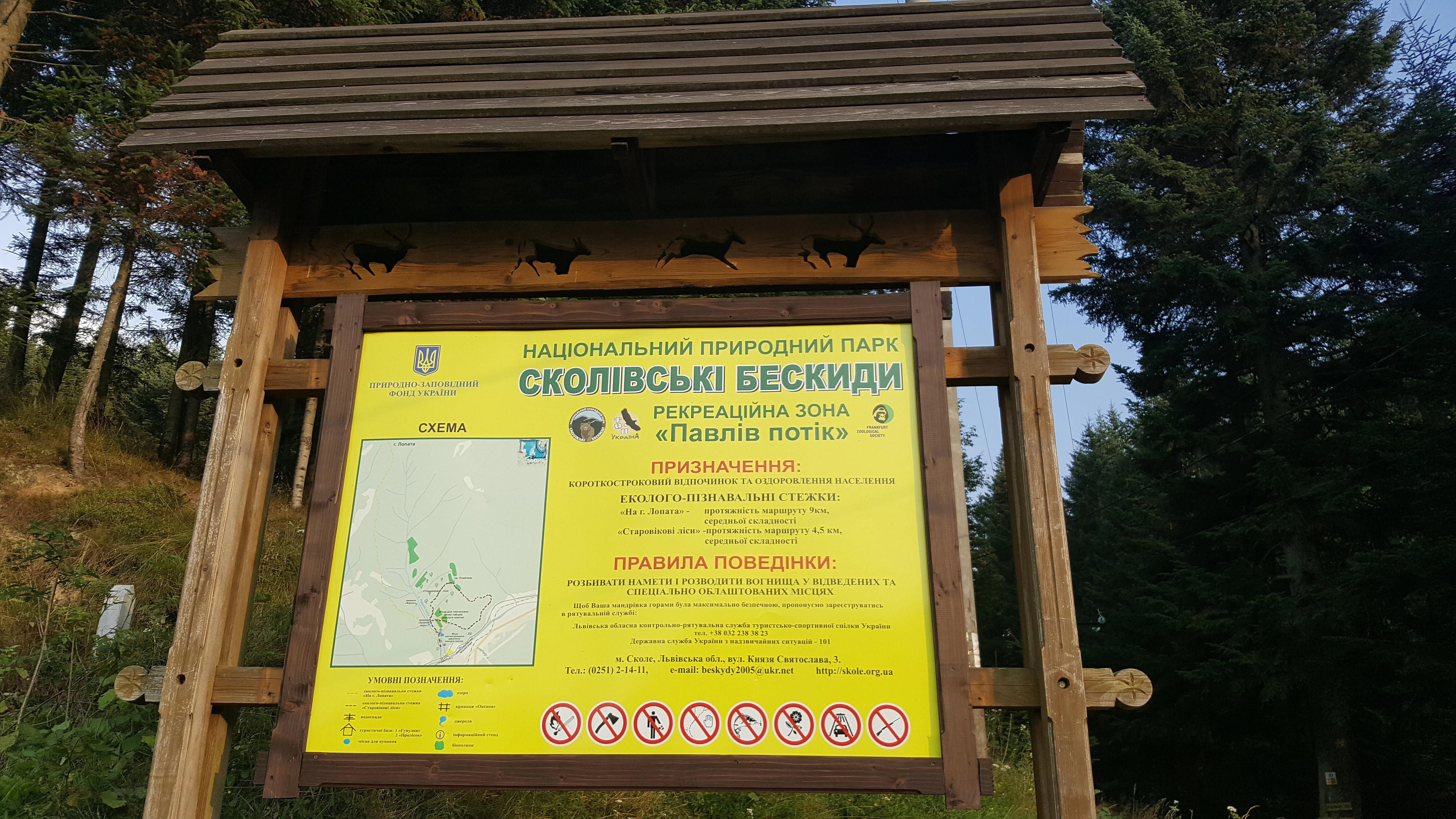 Wooden sign structure with yellow informational panel showing park name and map, located in a forested mountain area