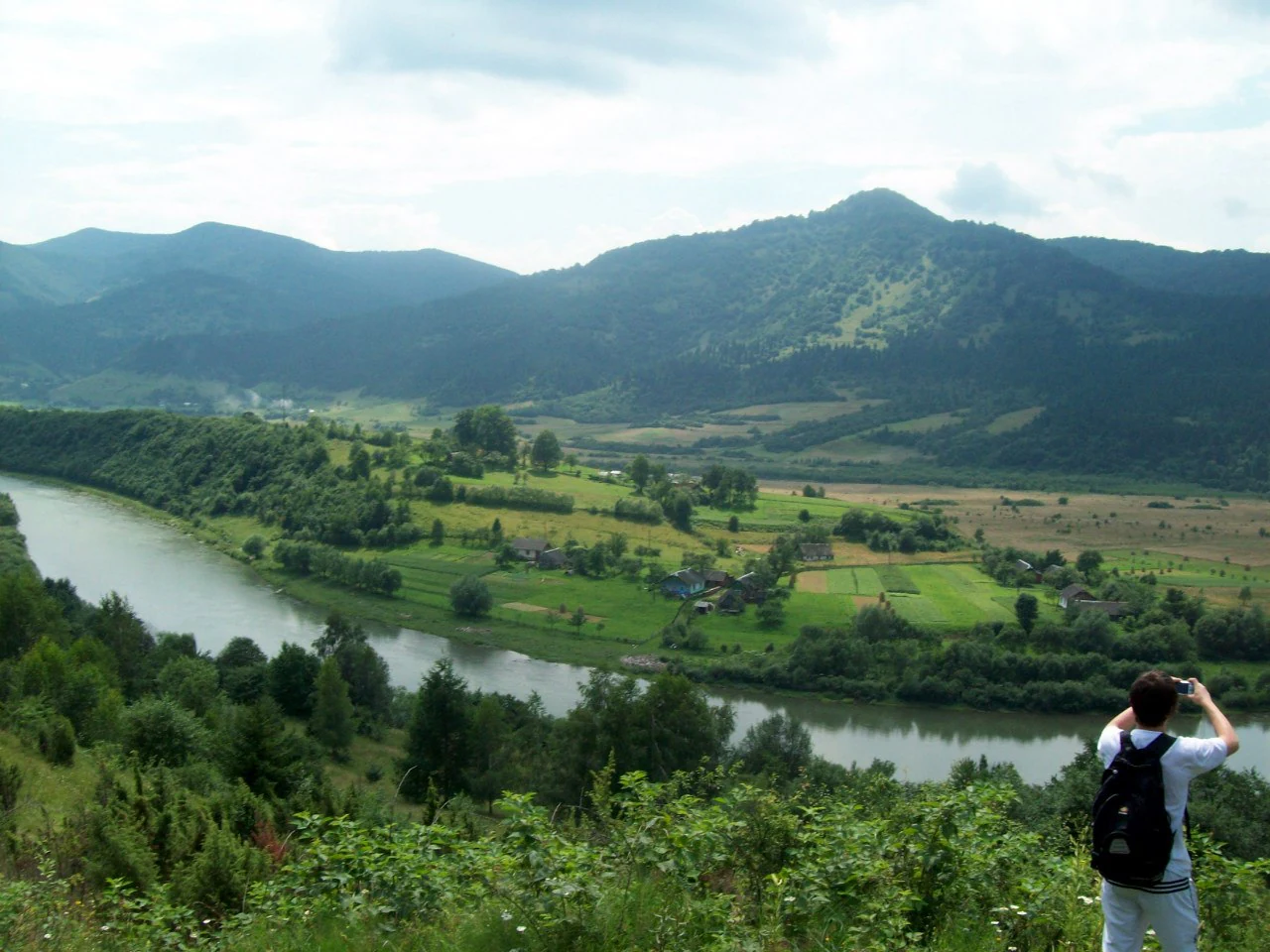Person with backpack standing at a viewpoint overlooking a river valley and mountain landscape