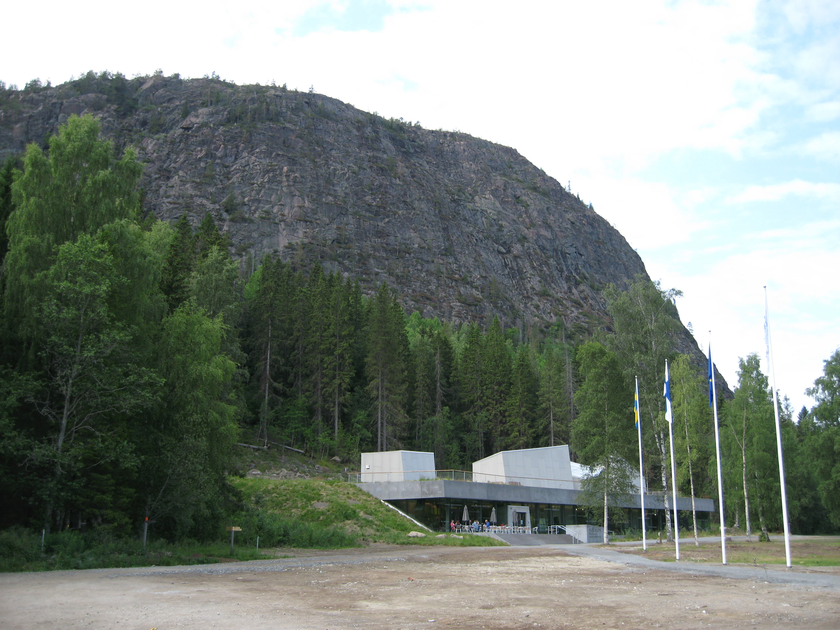 A large rocky mountain with a modern visitor center building and flagpoles in the foreground, surrounded by dense forest