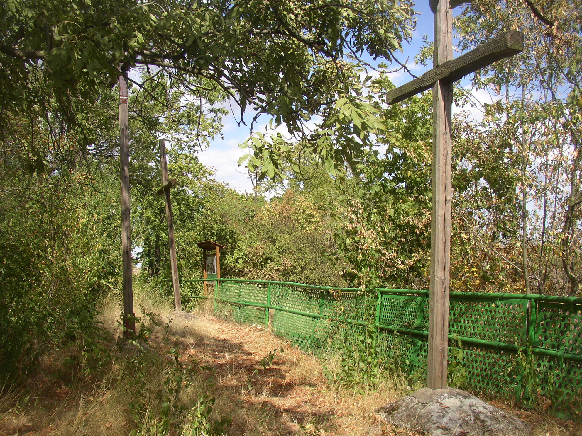 A dirt path along a wooded area featuring tall wooden crosses and a green metal fence with an educational trail sign in the background.