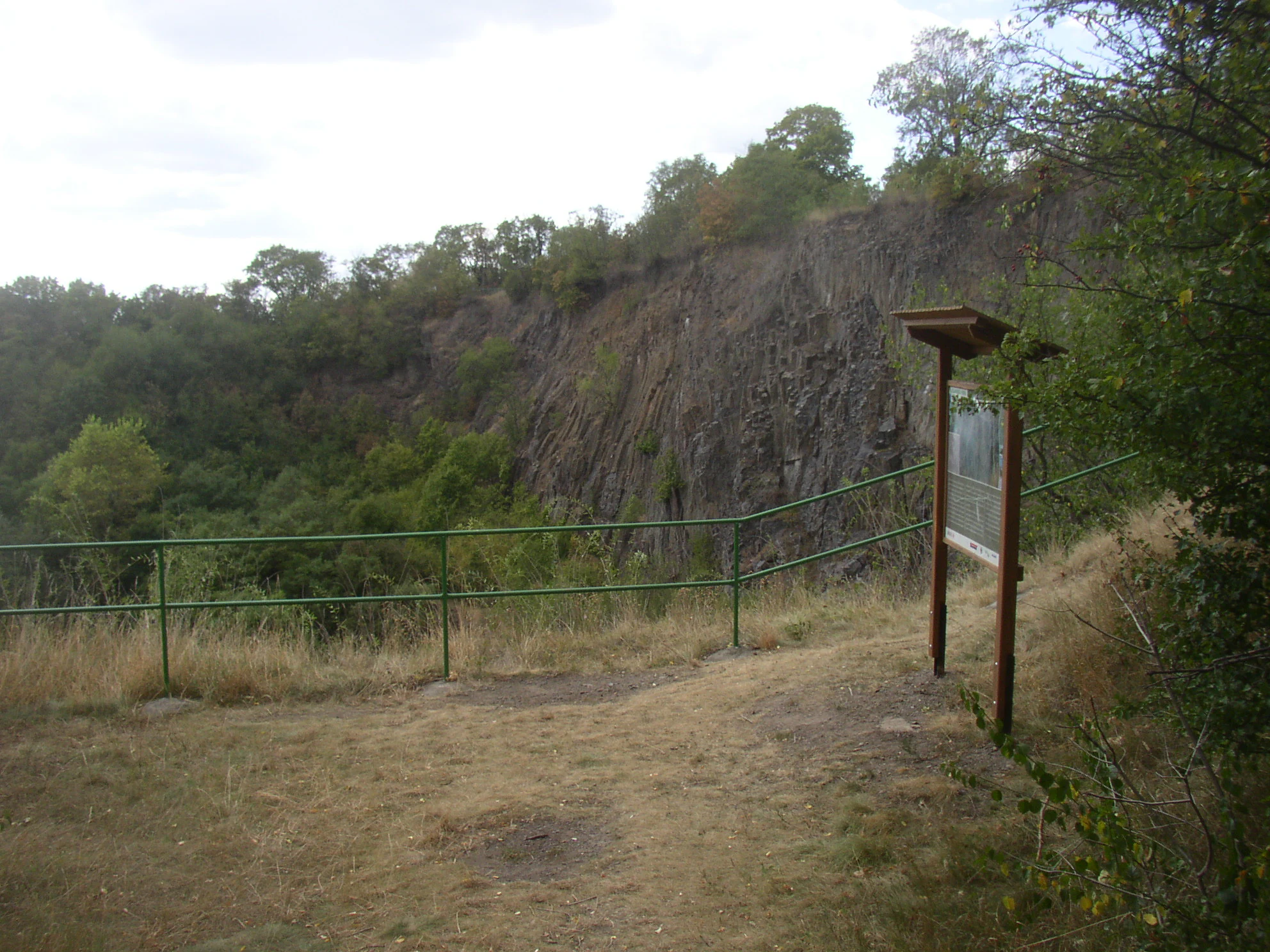 An educational information sign standing on a dirt path next to a green metal railing, overlooking a large volcanic rock cliff face covered in vegetation at Slánská hora.
