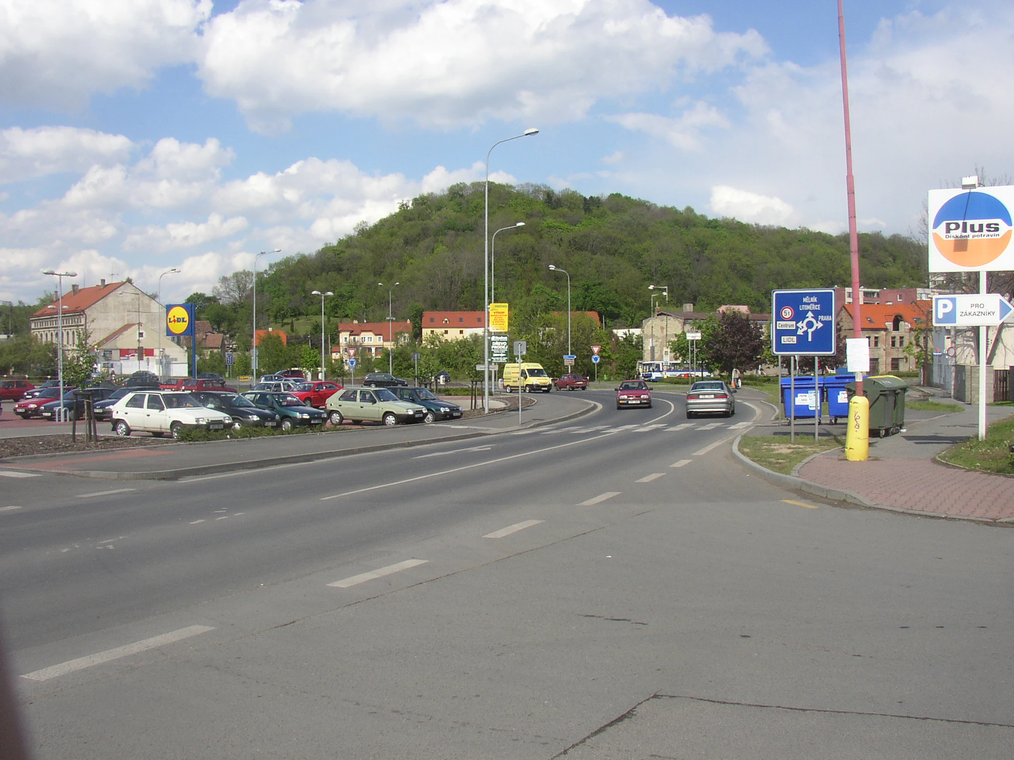A view of the forested Slánská hora hill rising above an urban road with parked cars, traffic signage, and commercial buildings.