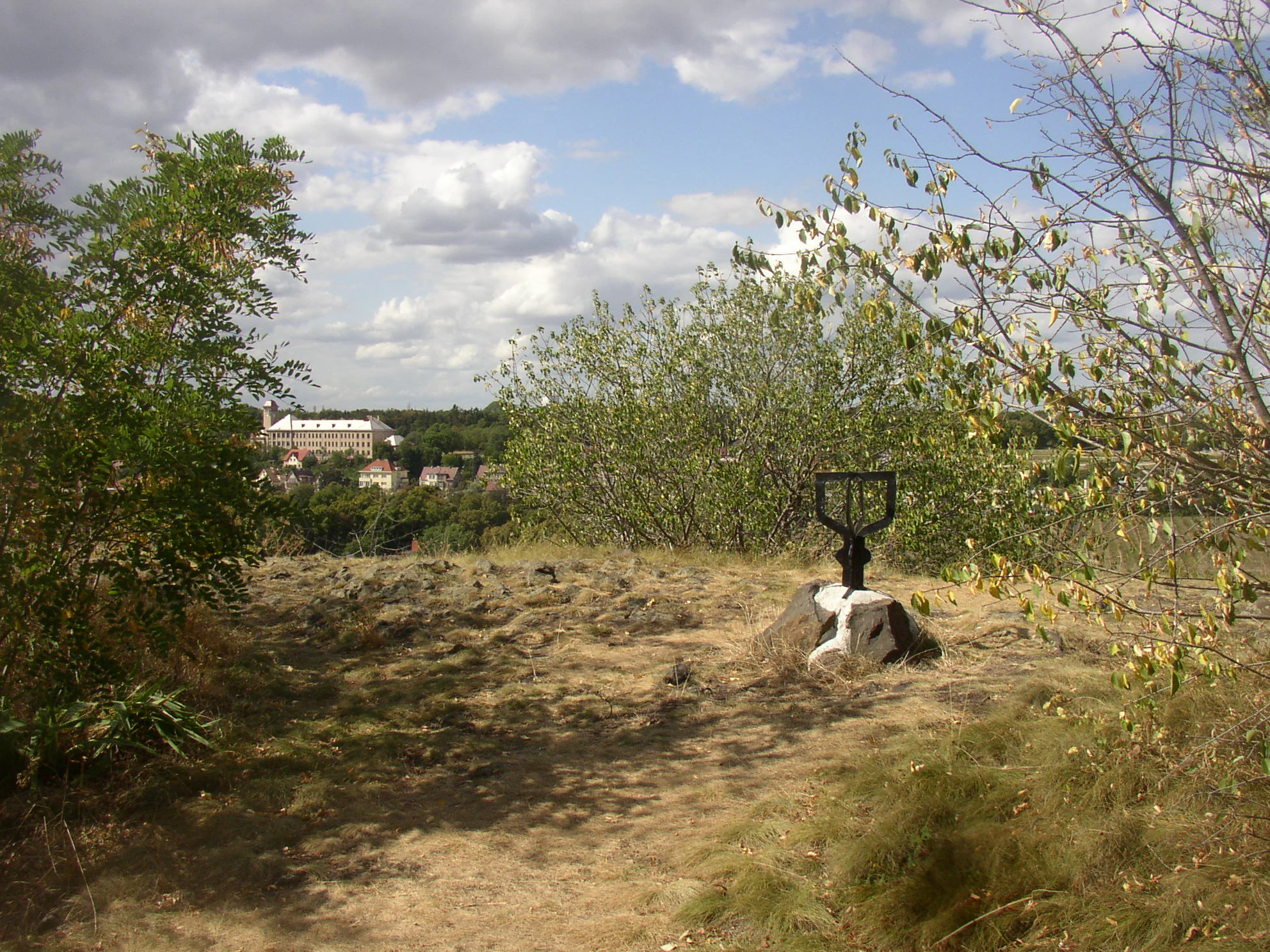 A dirt path leads to a summit area on a hill, featuring a small black metal marker mounted on a rock in the middle ground, with a distant town visible in the background.