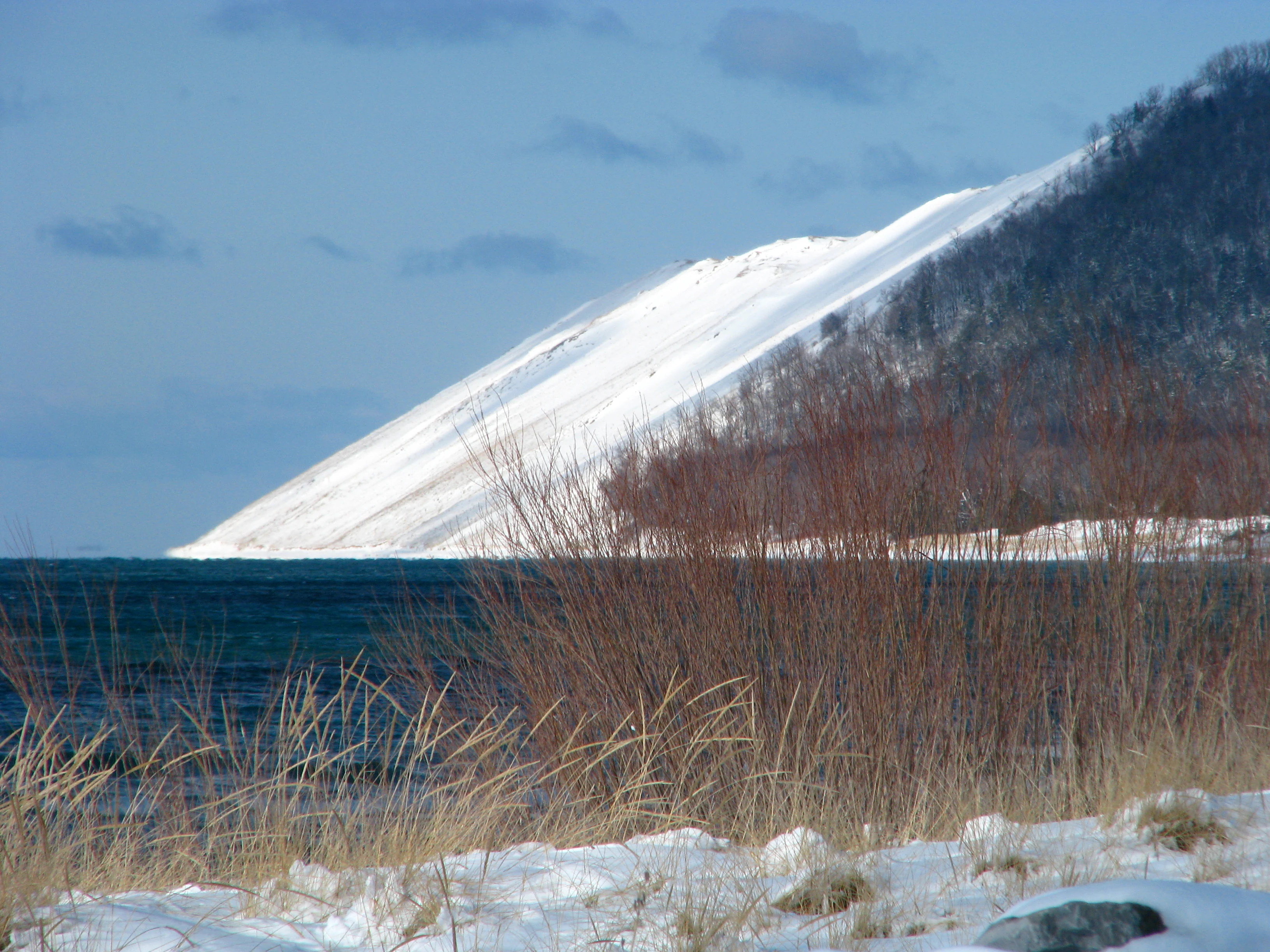 A steep, snow-covered sandy bluff rises sharply above the dark blue waters of Lake Michigan under a winter sky, with dried grasses visible in the foreground.