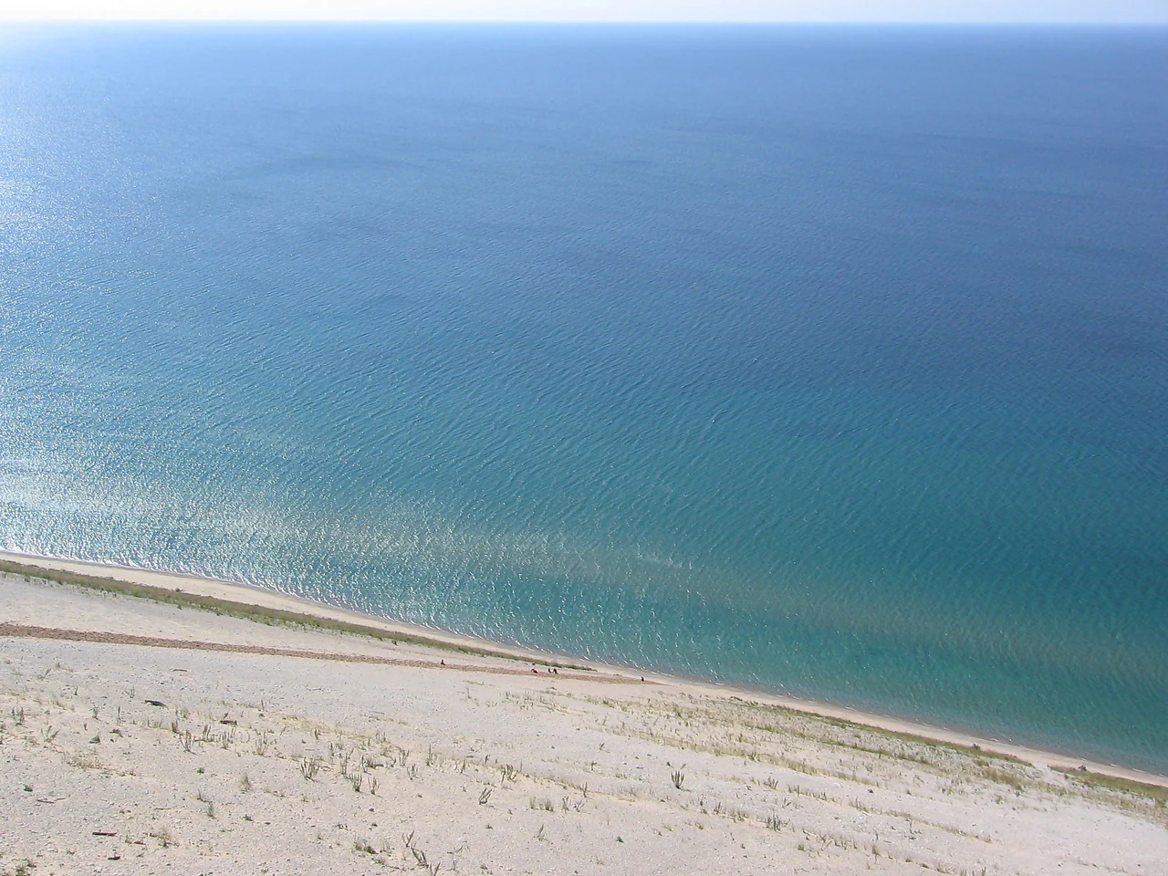 High angle view from a sandy bluff overlooking a long beach and the blue waters of Lake Michigan, with tiny silhouettes of hikers visible near the water's edge.