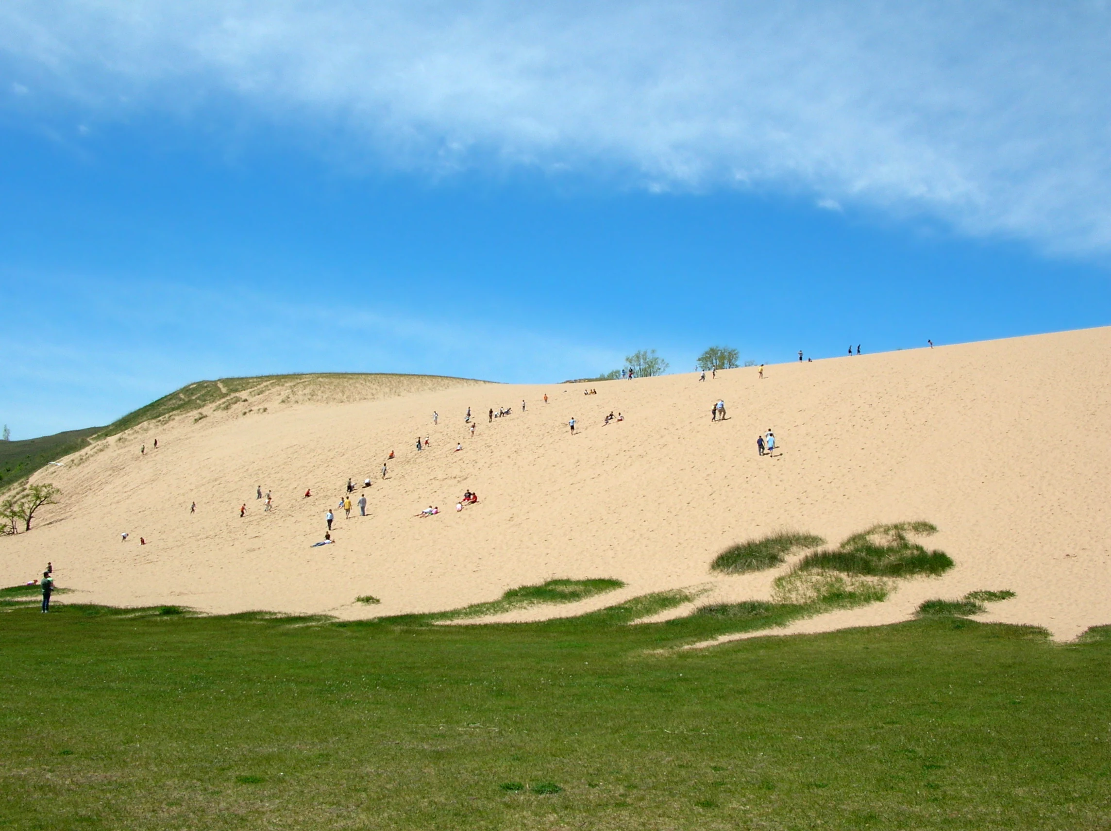 A wide, sunny shot of the Dune Climb at Sleeping Bear Dunes National Lakeshore, with many small figures of people ascending the steep, expansive sand slope from a green grassy base.