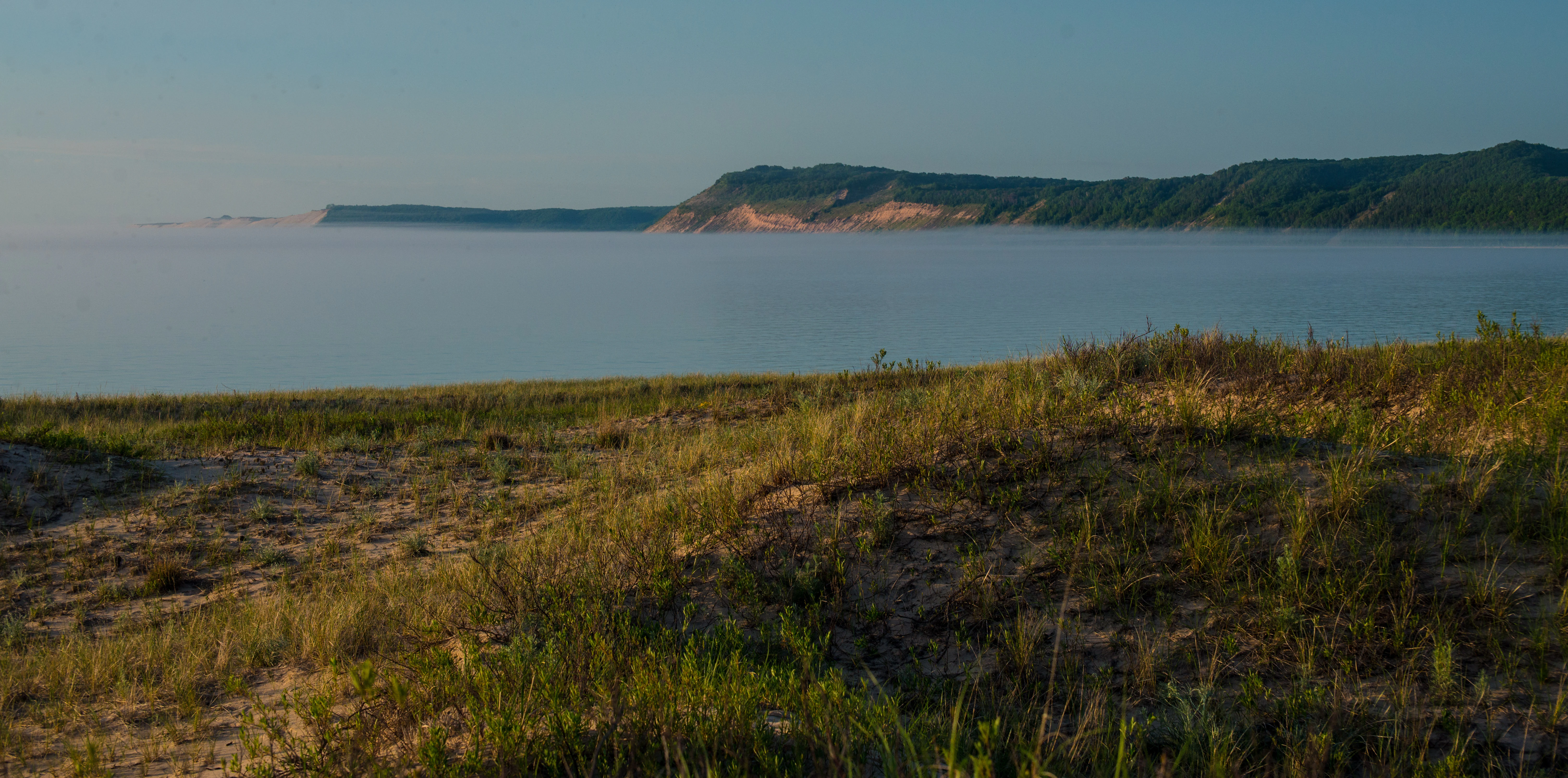 A low-angle view of grassy sand dunes in the foreground, looking out over a calm, foggy lake toward a distant, forested ridge line under a clear sky.