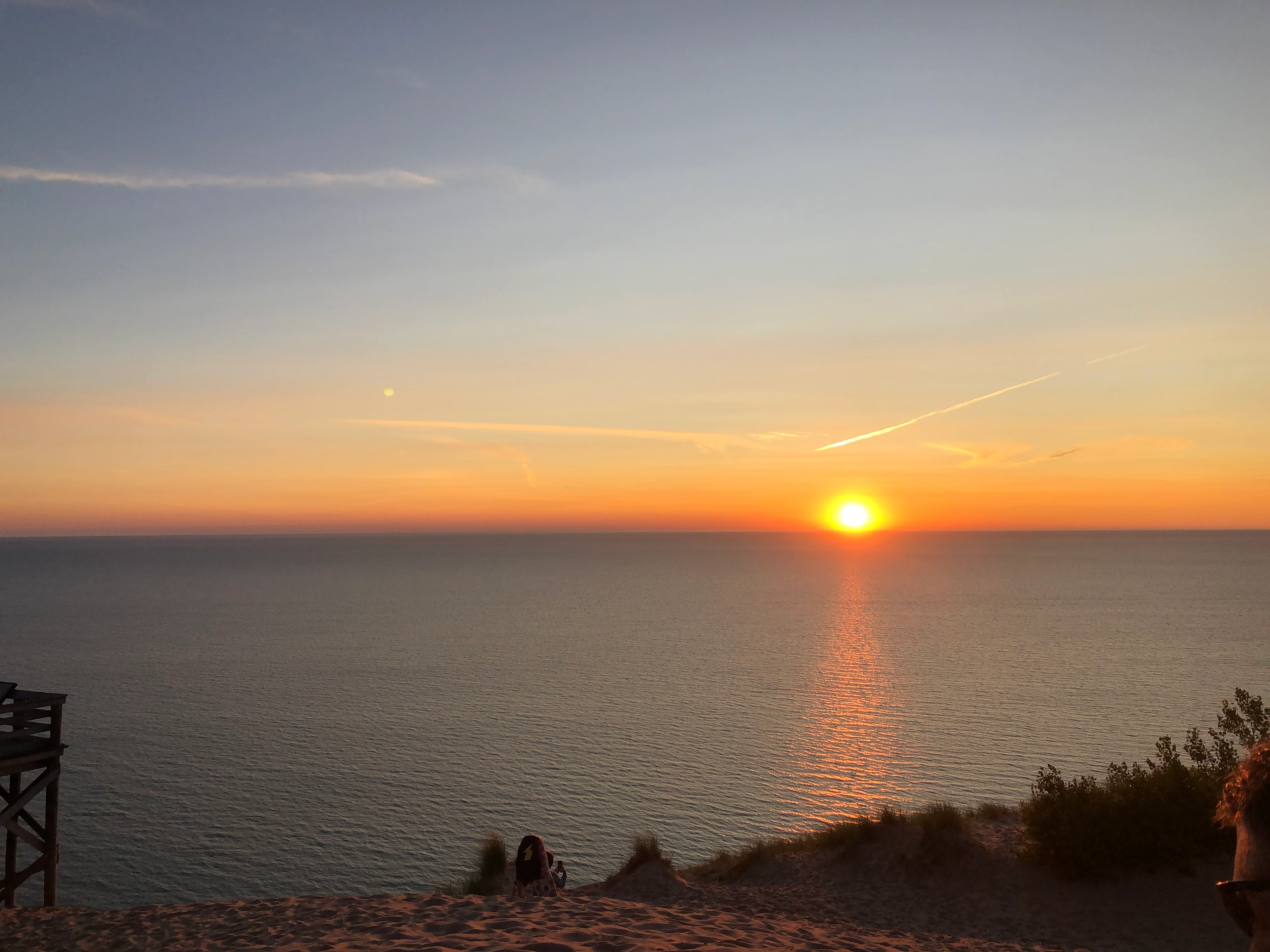 The sun sets over the horizon of a large lake, casting a golden glow on the water; in the foreground are sand dunes with a wooden observation structure visible on the left.