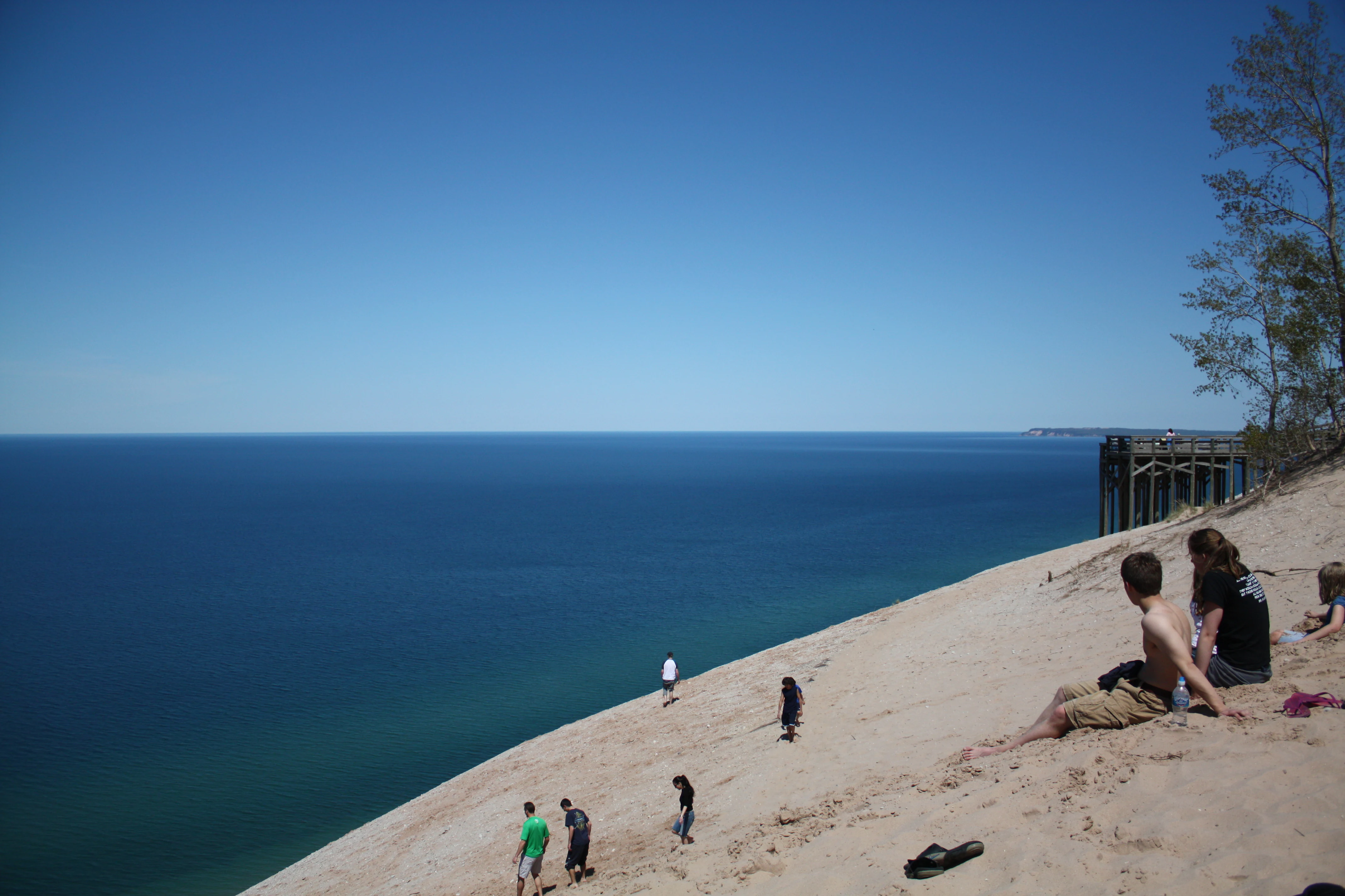 A group of people sitting or standing on the steep, sandy slope of a large dune overlooking the deep blue waters of Lake Michigan.