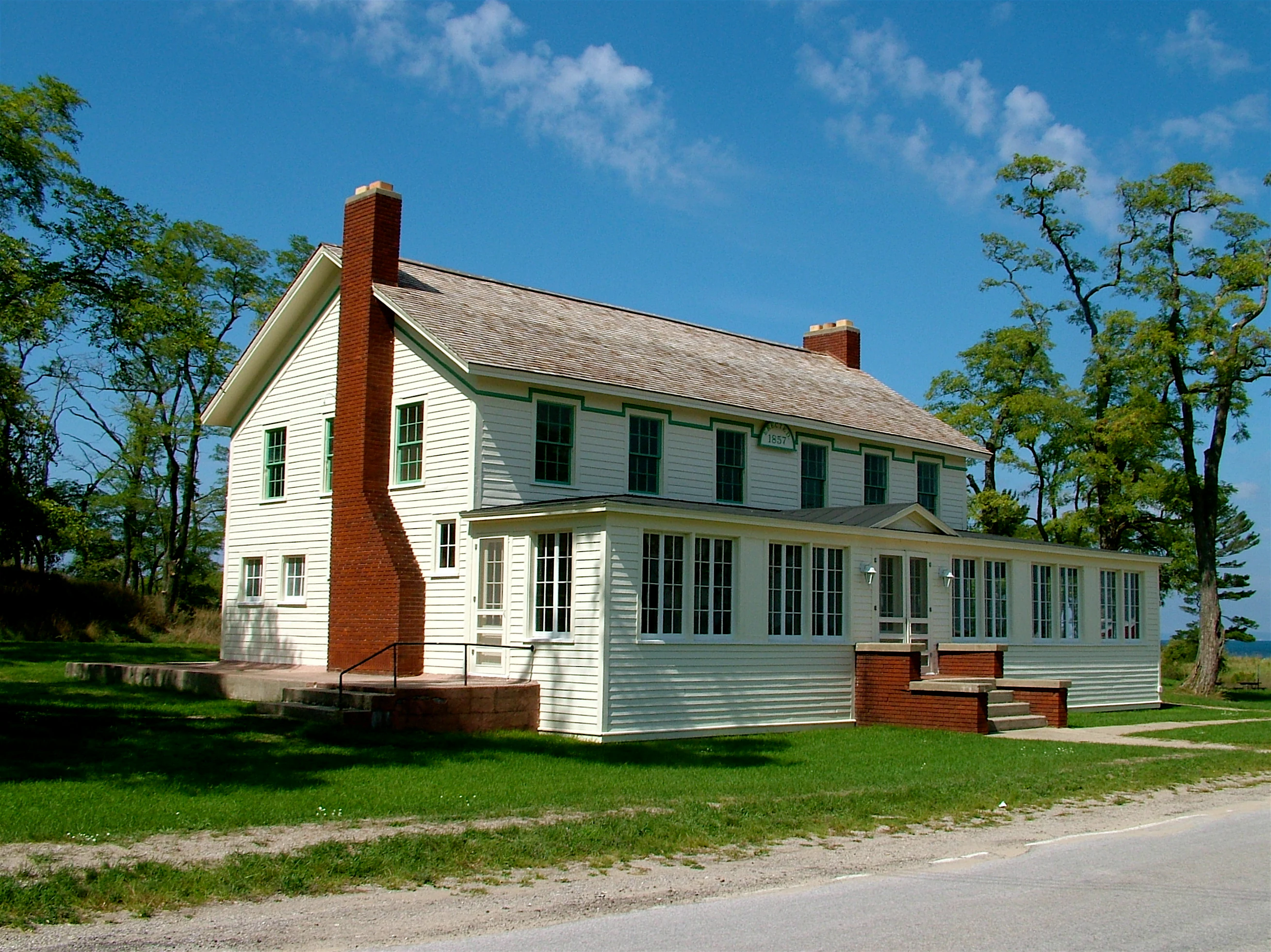 A two-story, white-sided historic inn with a prominent red brick chimney and a long front porch, situated on a grassy lawn with trees and a clear blue sky.