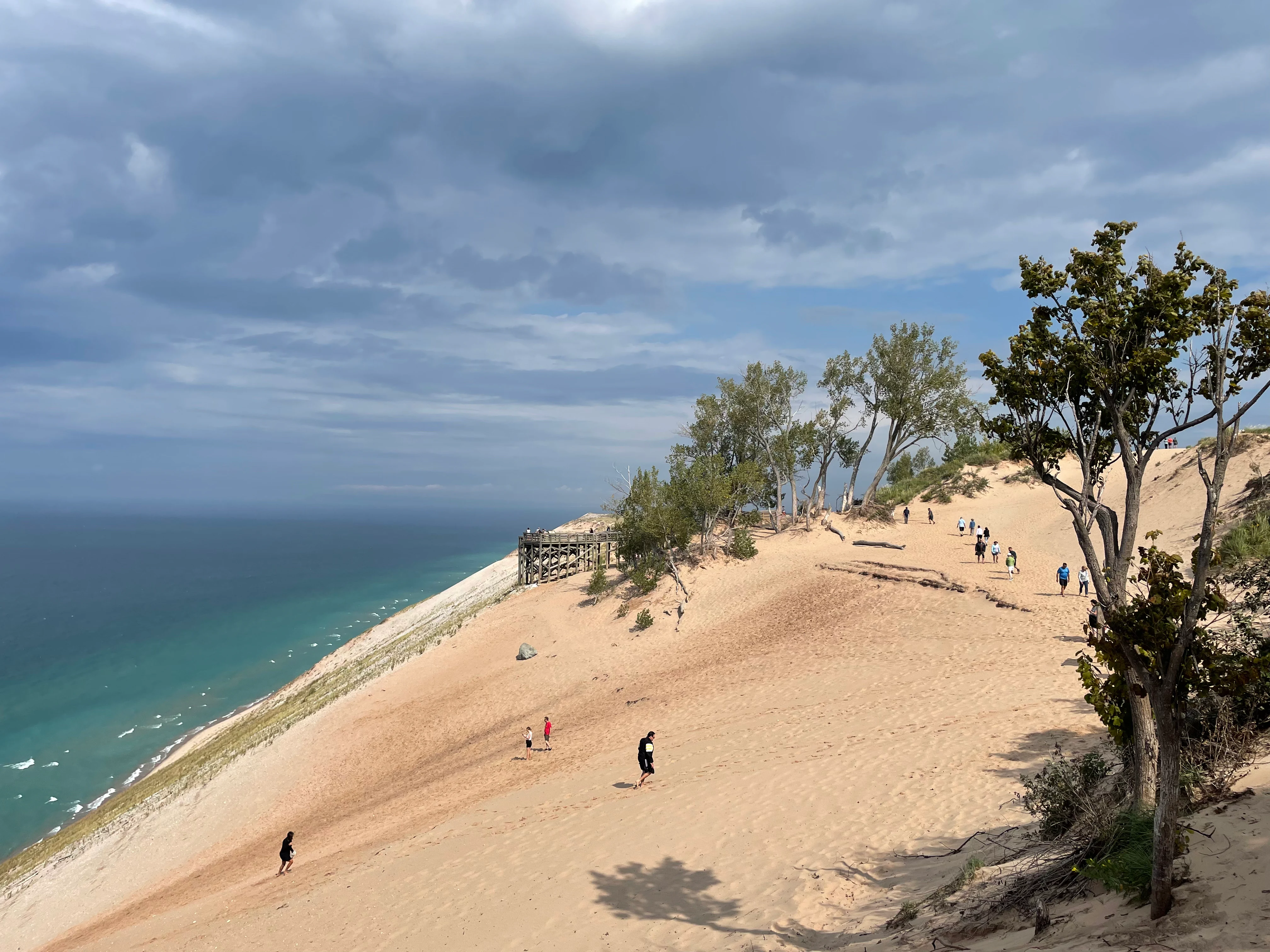 A wide view of the Sleeping Bear Dunes, featuring large, steep sand slopes, sparse trees, and a wooden viewing platform, with people walking on the sand and the blue water of Lake Michigan in the distance under a cloudy sky.