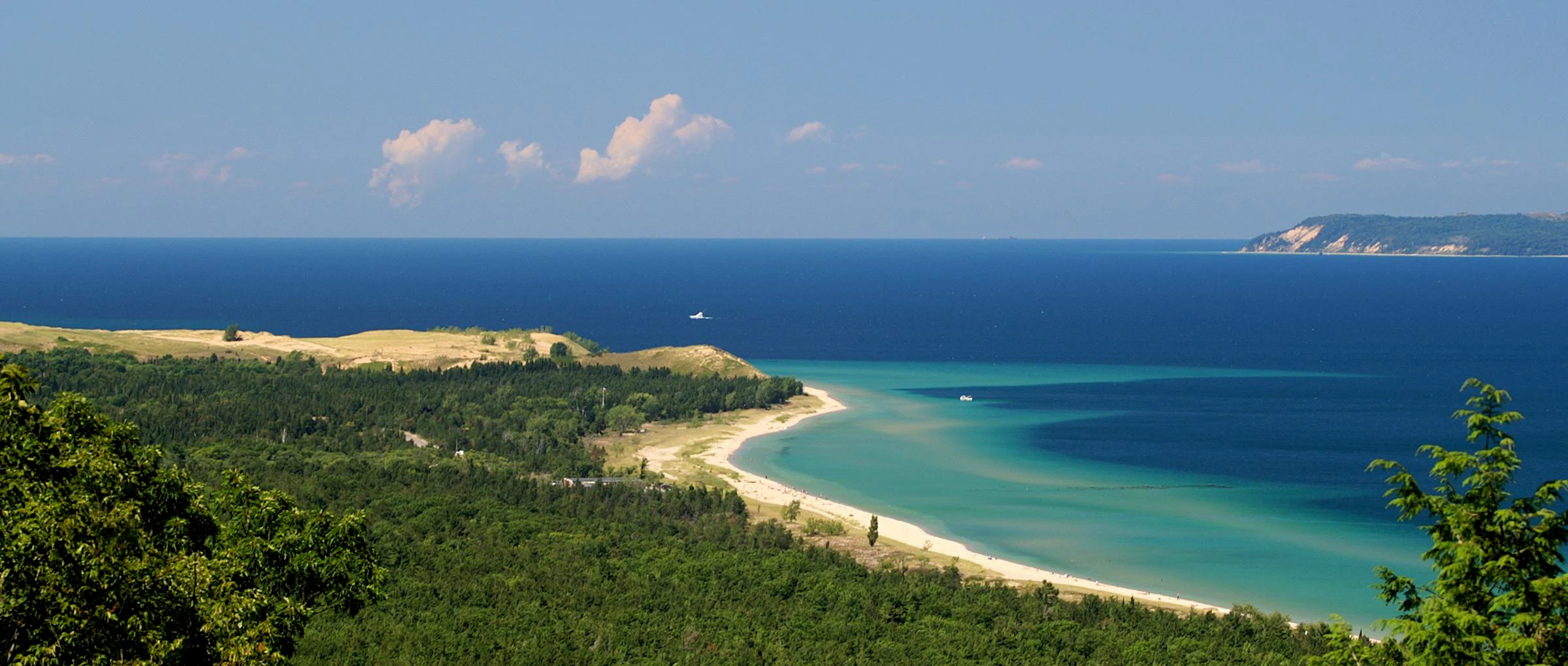 An elevated, wide-angle view of a sandy beach curving along a forested coastline, meeting the deep blue waters of Lake Michigan under a clear sky.