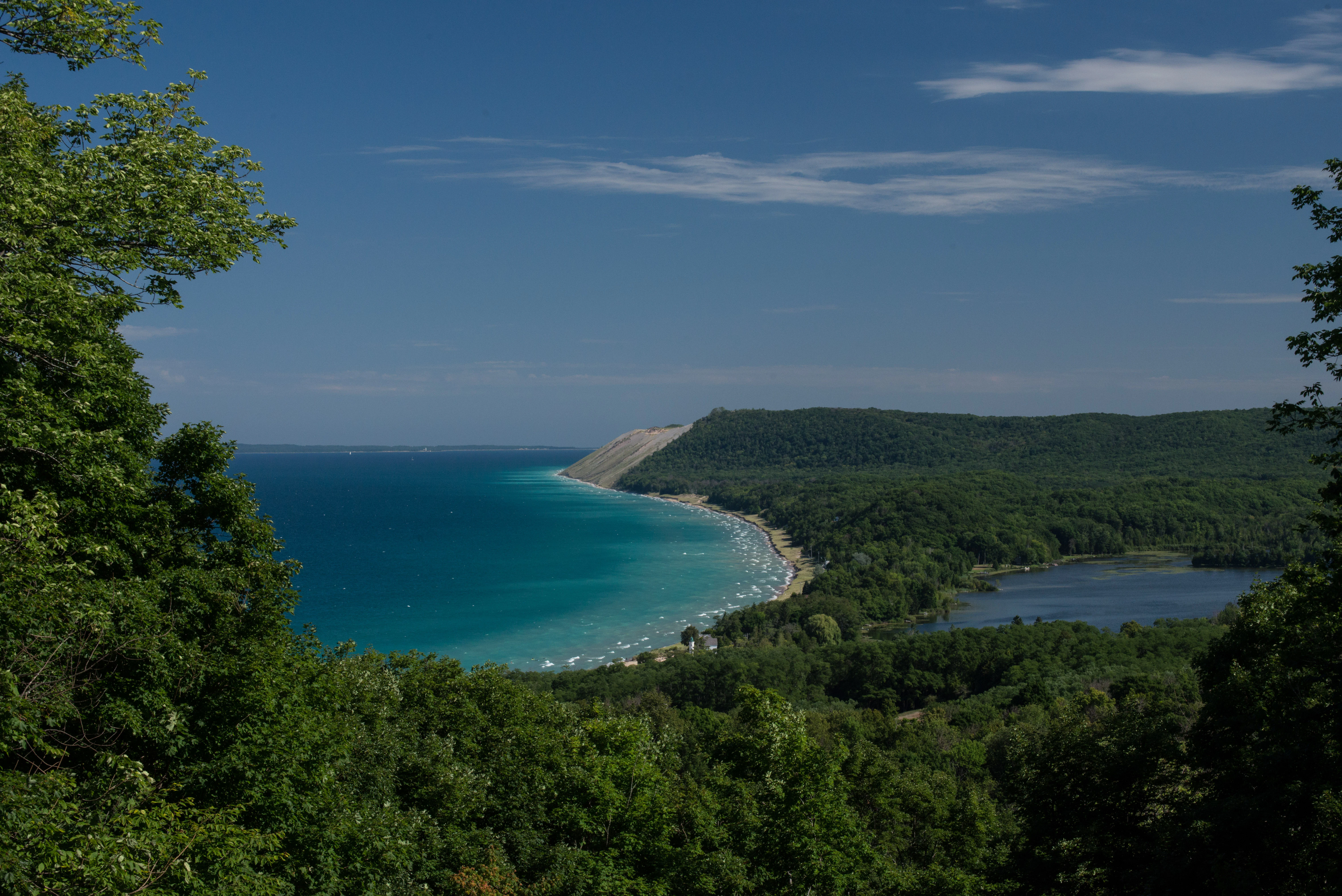 A wide angle view from a forested bluff overlooking a blue Lake Michigan shoreline, sandy dunes in the distance, and a small inland lake nestled in the green forest.