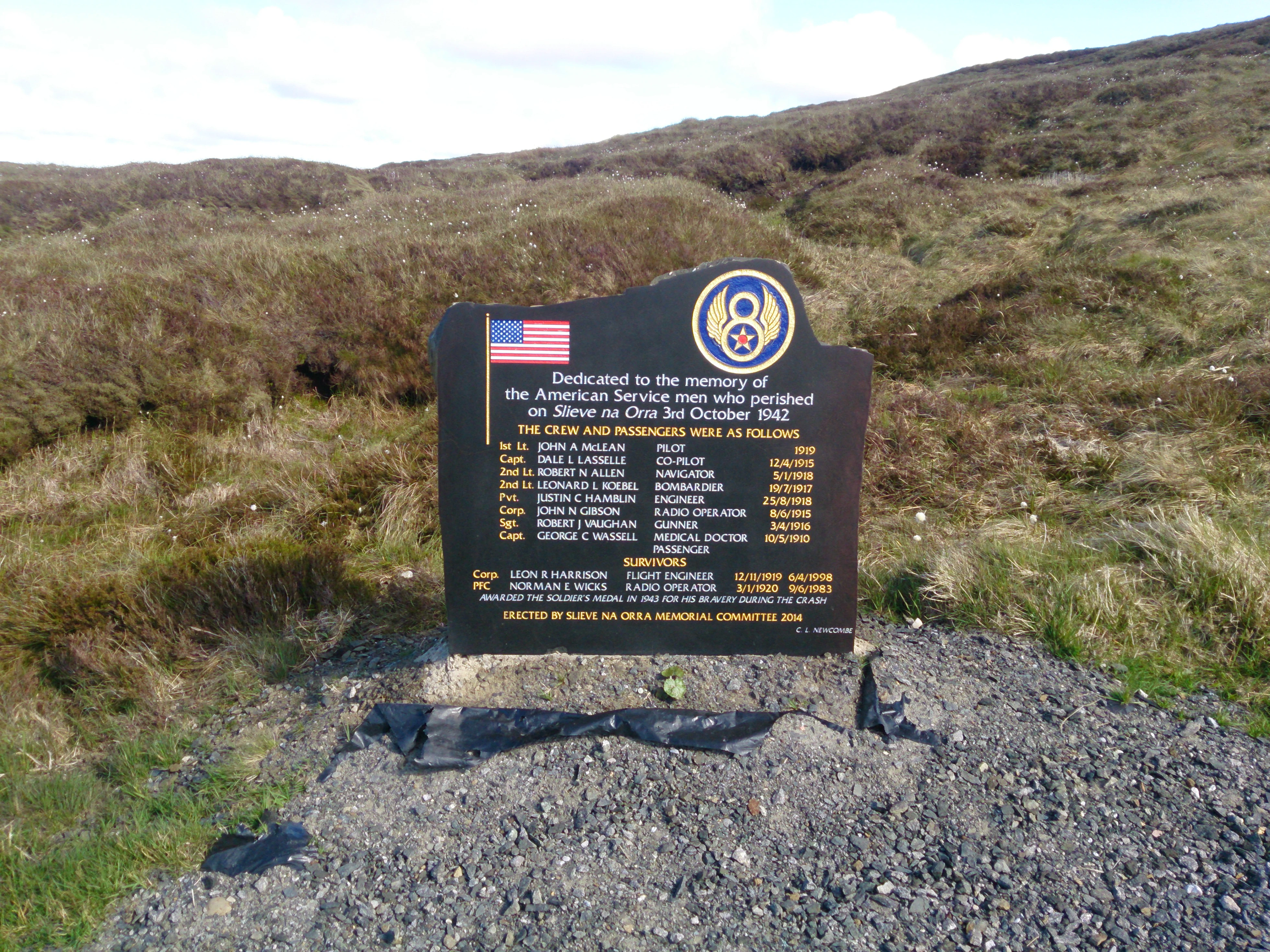 Una placa conmemorativa negra con una bandera estadounidense y el emblema de la Octava Fuerza Aérea que enumera los nombres de los militares; se encuentra sobre una base de grava en un paisaje herboso y montañoso.