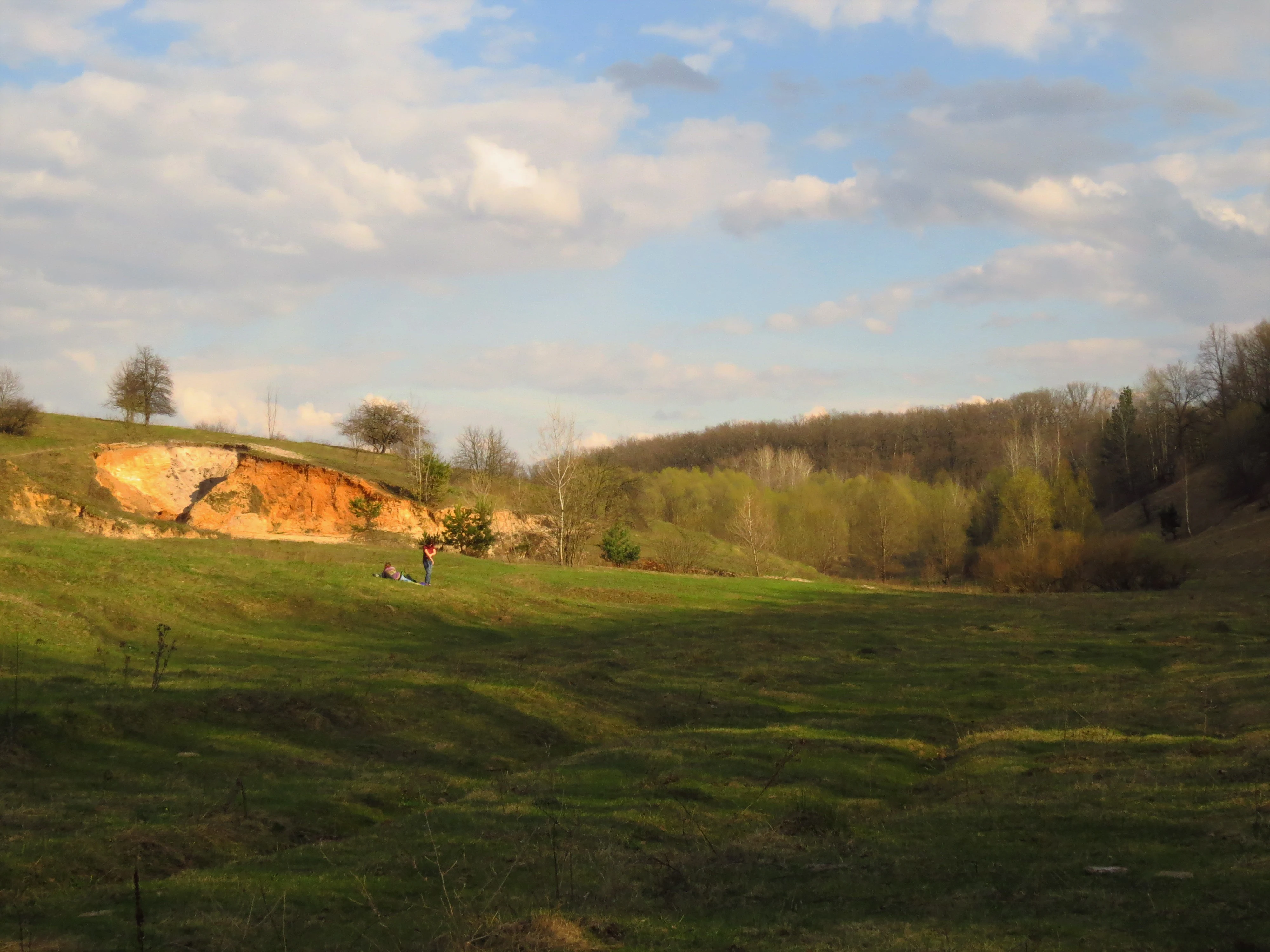 Grassy field with a reddish-brown clay cliff, scattered trees, and a few people walking in the distance under a partly cloudy sky