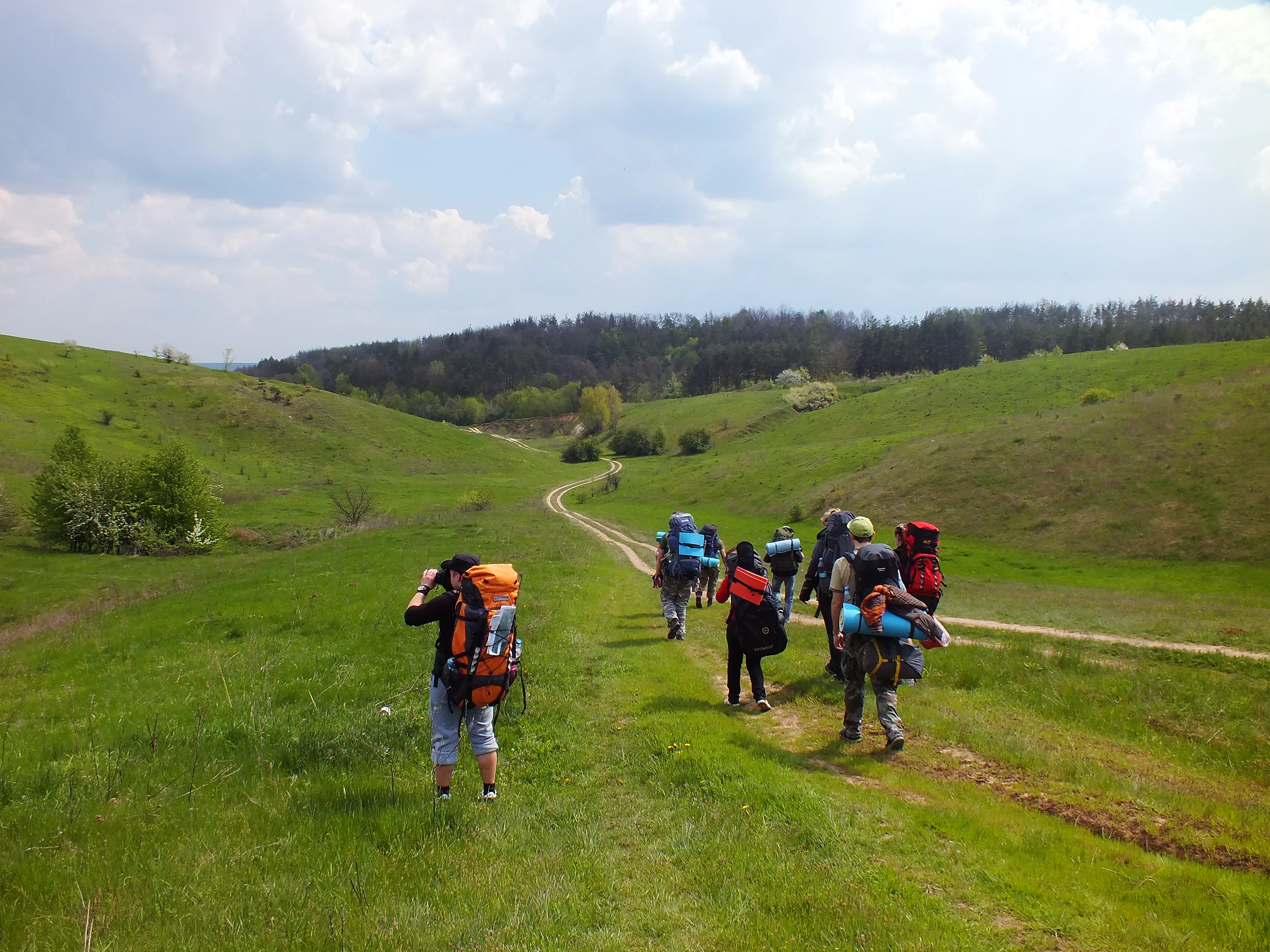 Group of hikers with backpacks walking on a dirt path through a grassy landscape with hills in the background