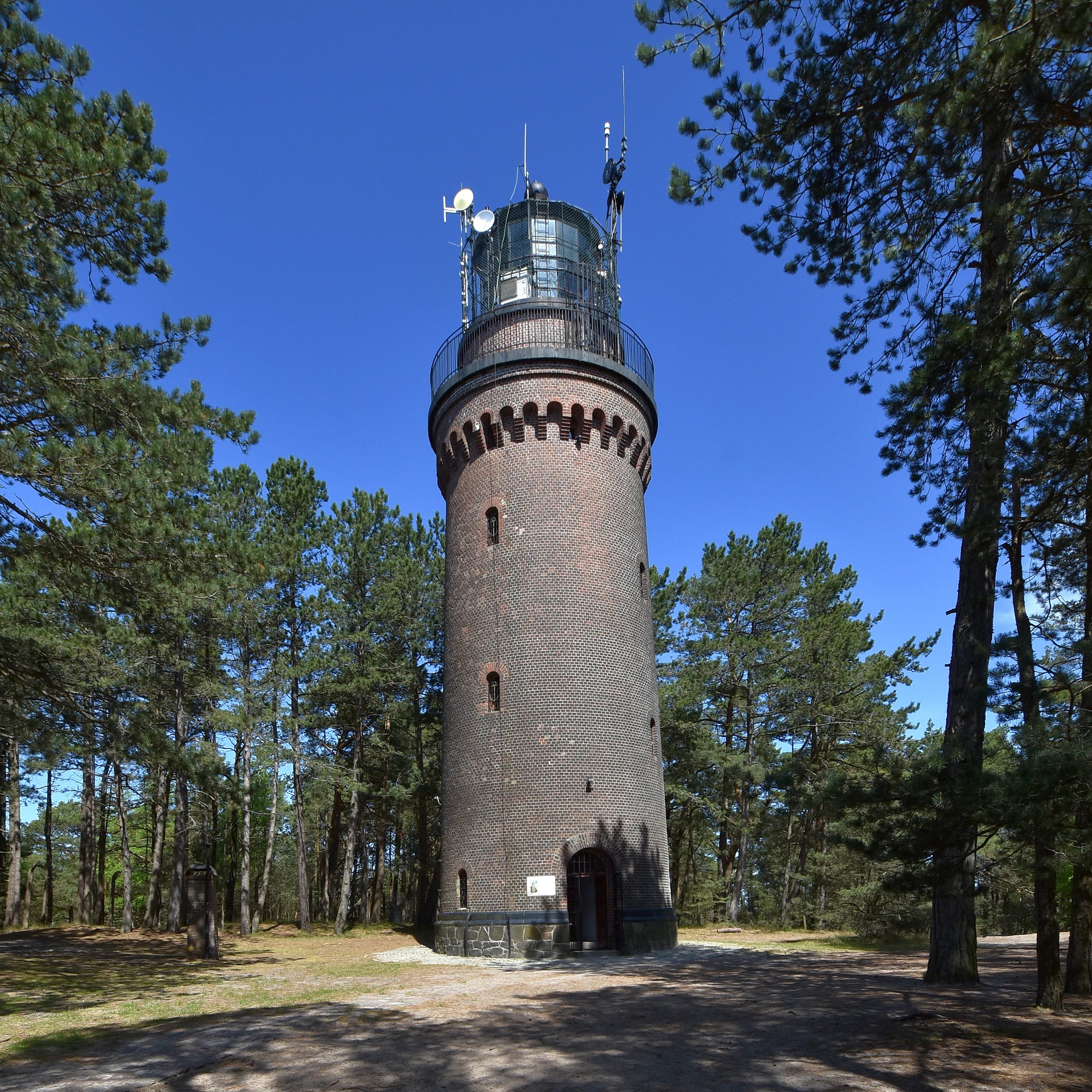 A tall, red-brick lighthouse with a black lantern room at the top, surrounded by pine trees under a clear blue sky.