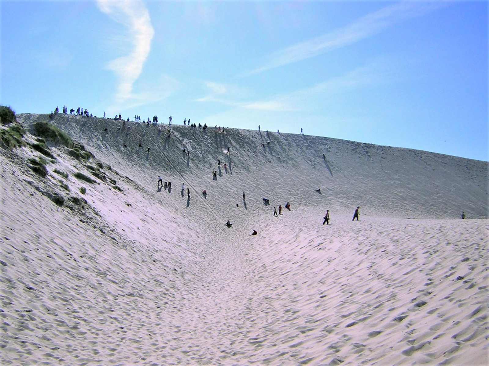 A wide, sunny view of a large, steep sand dune with many small figures of people walking up the slope against a clear blue sky.