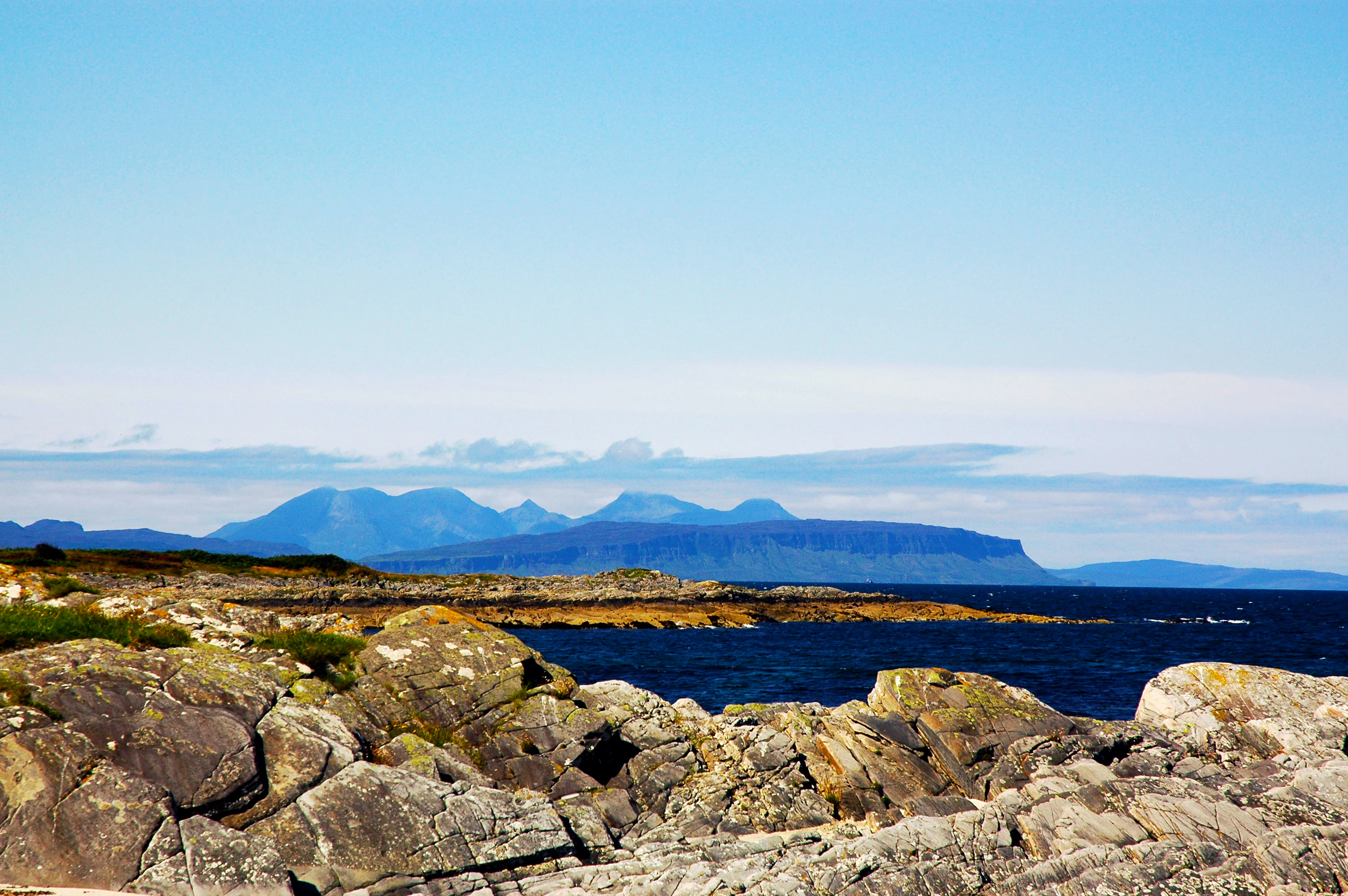 A coastal landscape looking across a rocky foreground and blue sea toward a series of distant, hazy mountain islands on the horizon.