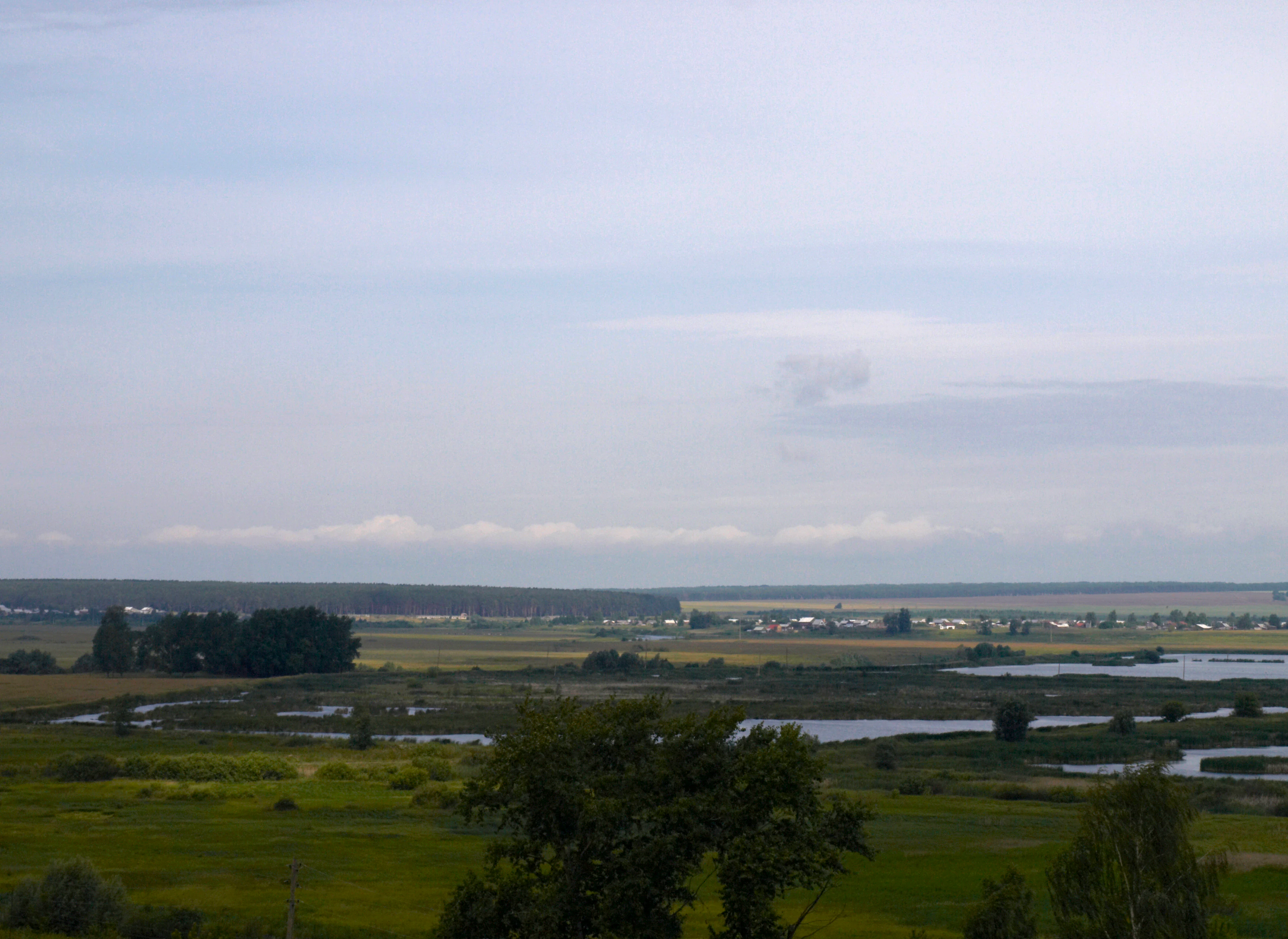 Wide landscape view showing the Alatyr River flowing through wetlands with fields, trees, and distant settlements under a partly cloudy sky