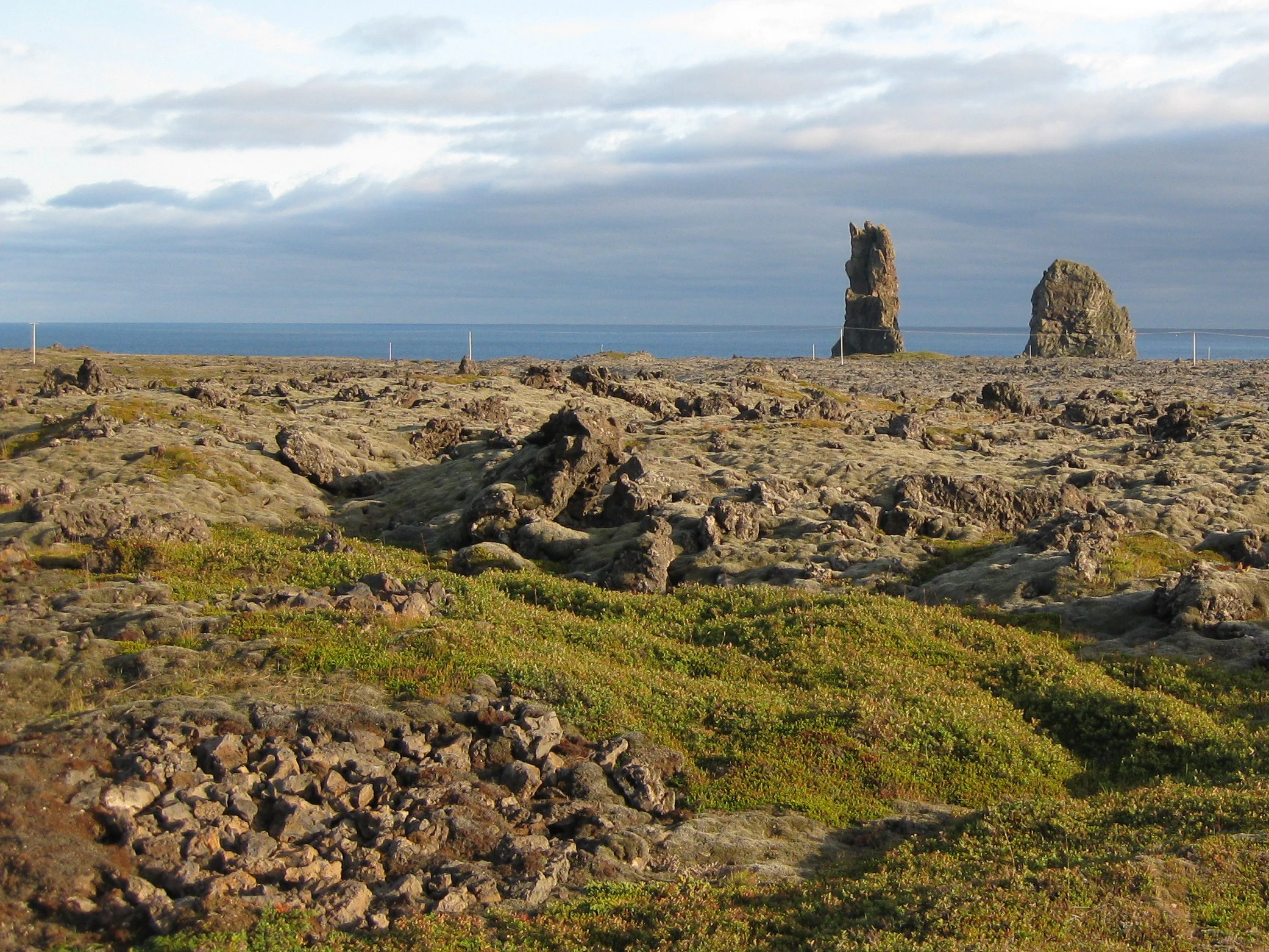 Una vista amplia de un campo de lava rocoso y cubierto de musgo que se extiende hacia dos grandes pilares de basalto irregulares situados cerca del océano bajo un cielo nublado.
