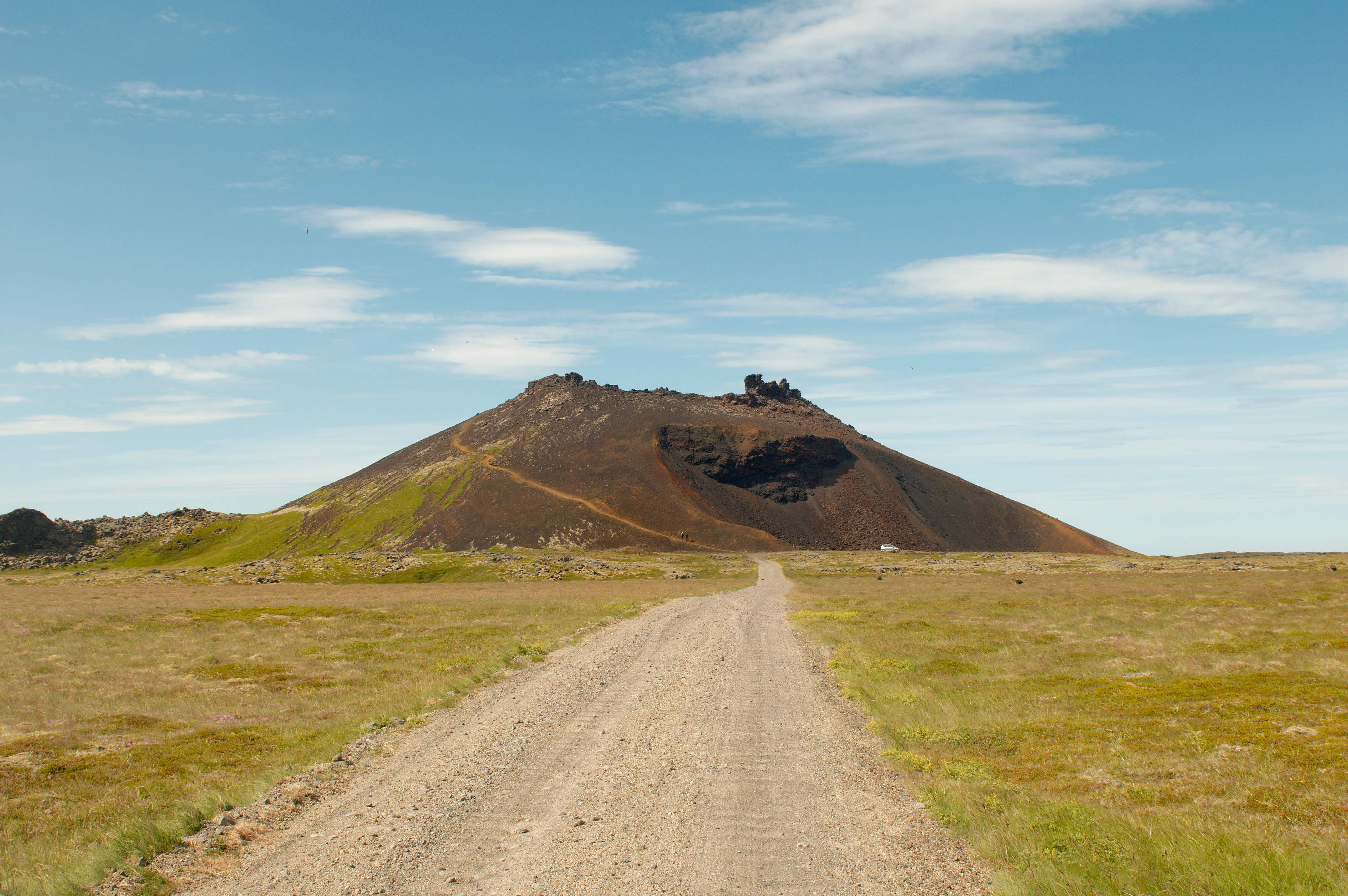 Un camino de tierra ancho se extiende a través de un paisaje volcánico plano y cubierto de hierba hacia un prominente cráter volcánico de color oscuro bajo un cielo parcialmente nublado.
