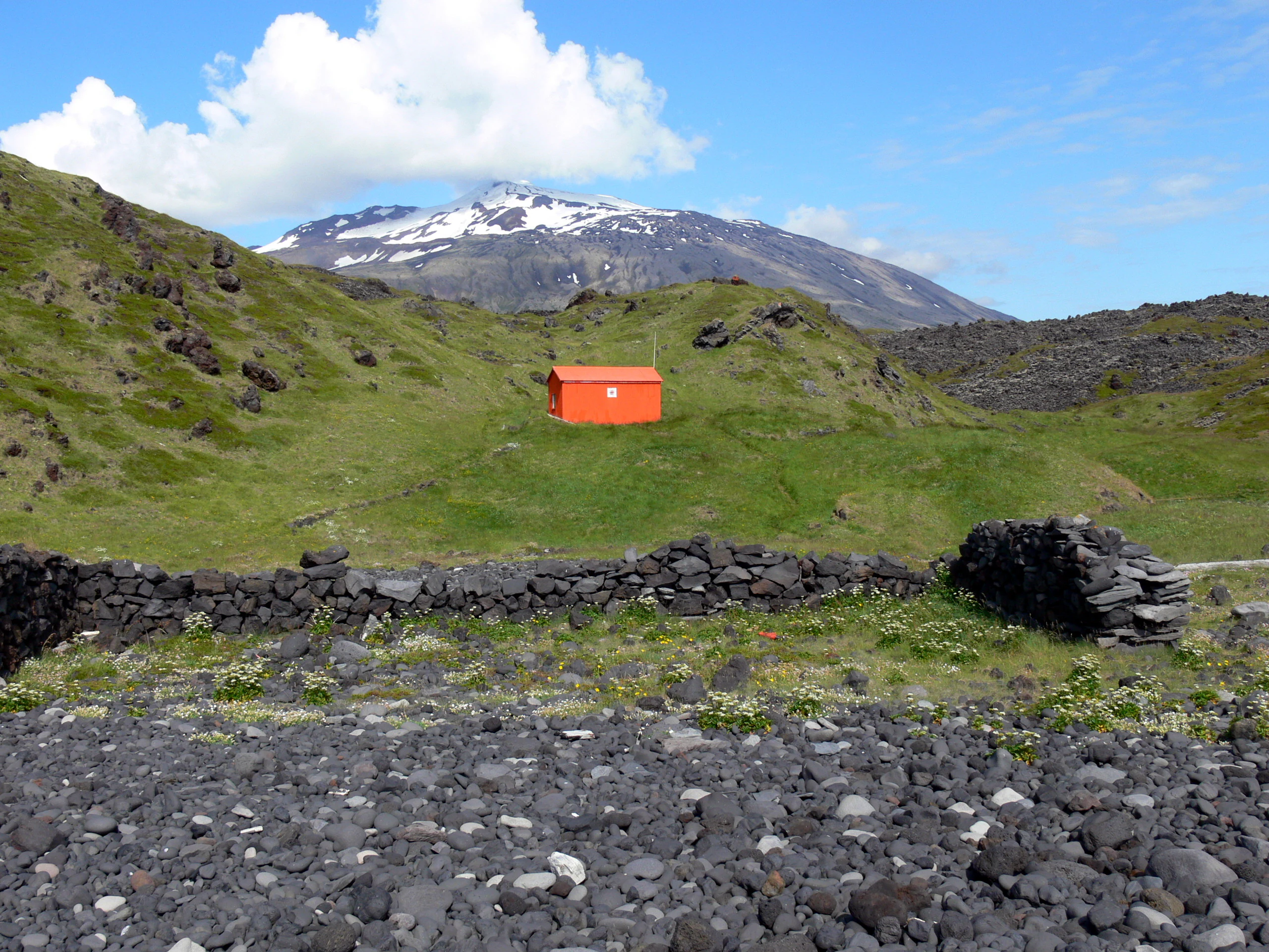 Una vista amplia de un campo de rocas volcánicas oscuras en primer plano, que conduce a una pequeña estructura naranja situada entre colinas verdes, con un pico montañoso nevado que se alza al fondo bajo un cielo azul.