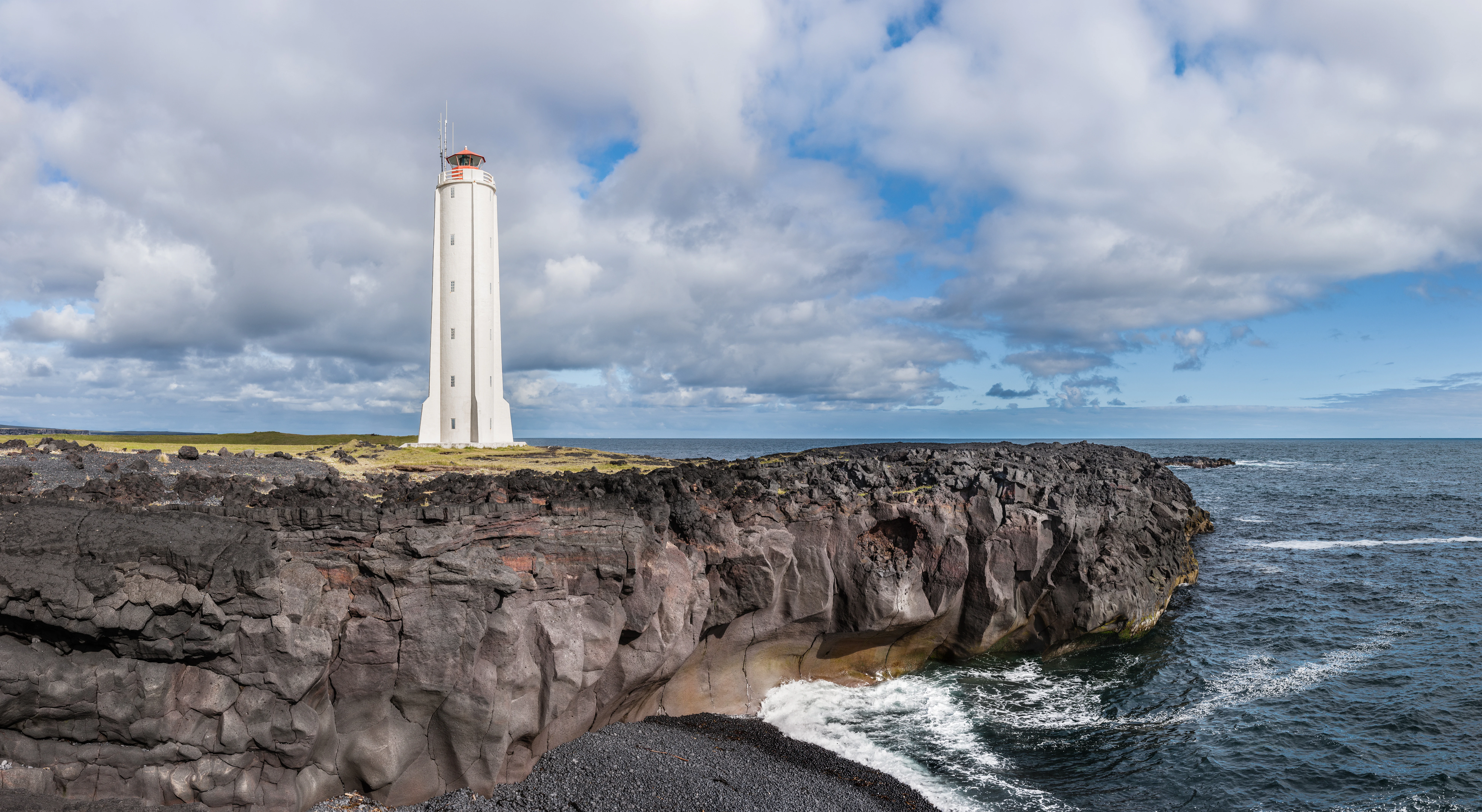 Un faro blanco con la parte superior roja se alza sobre un acantilado de roca volcánica oscura y escarpada, con vistas al océano bajo un cielo nublado.
