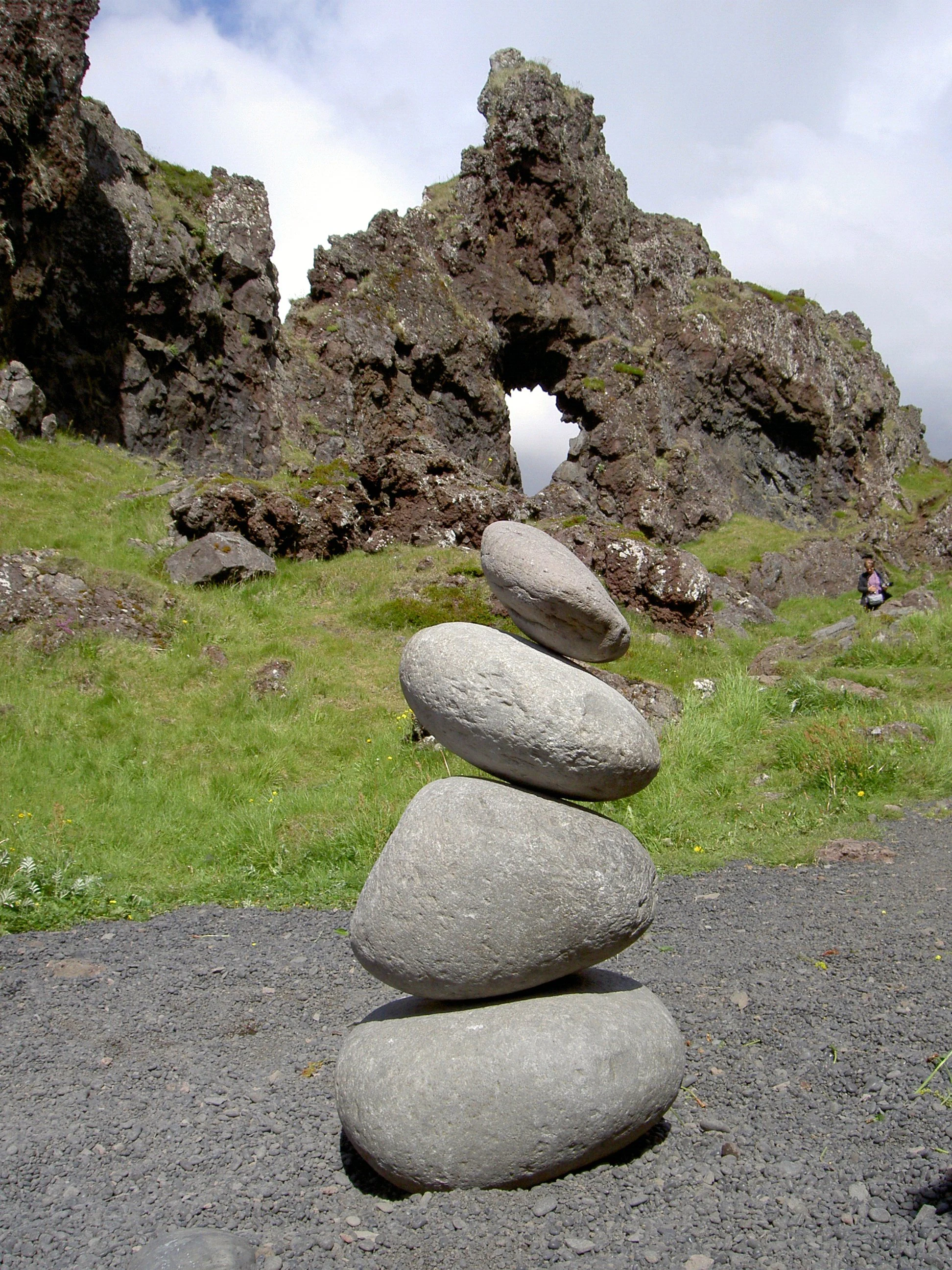 Una pila de cuatro piedras grises grandes, redondeadas y lisas, colocadas verticalmente sobre un sendero de grava oscura, con un acantilado volcánico rocoso que presenta un arco natural al fondo.