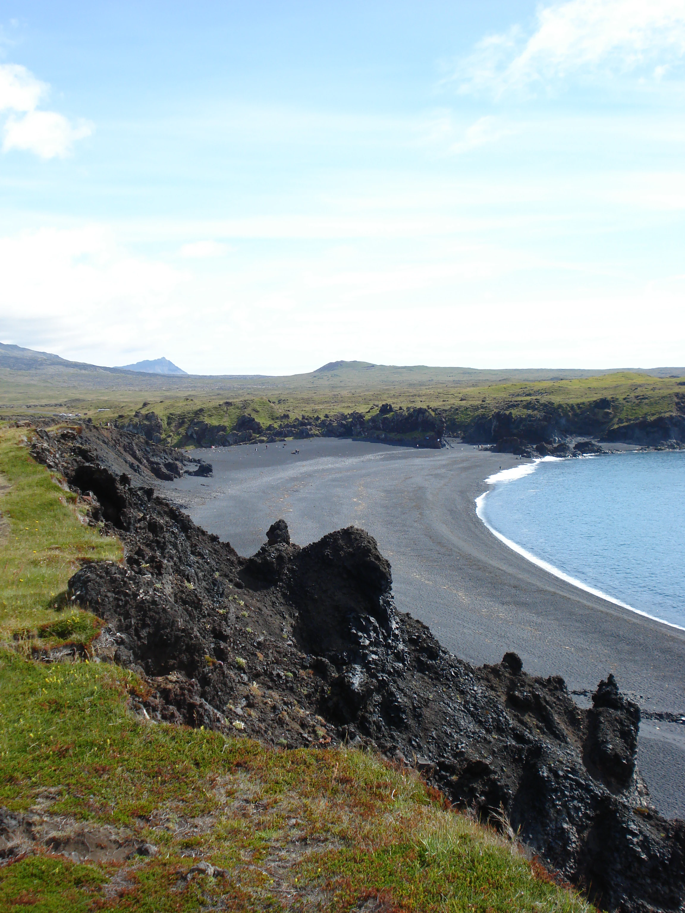 Vista en ángulo elevado de una playa de arena negra y guijarros en forma de media luna, bordeada por formaciones rocosas volcánicas escarpadas, con una ensenada de océano azul y tundra costera verde al fondo bajo un cielo parcialmente nublado.