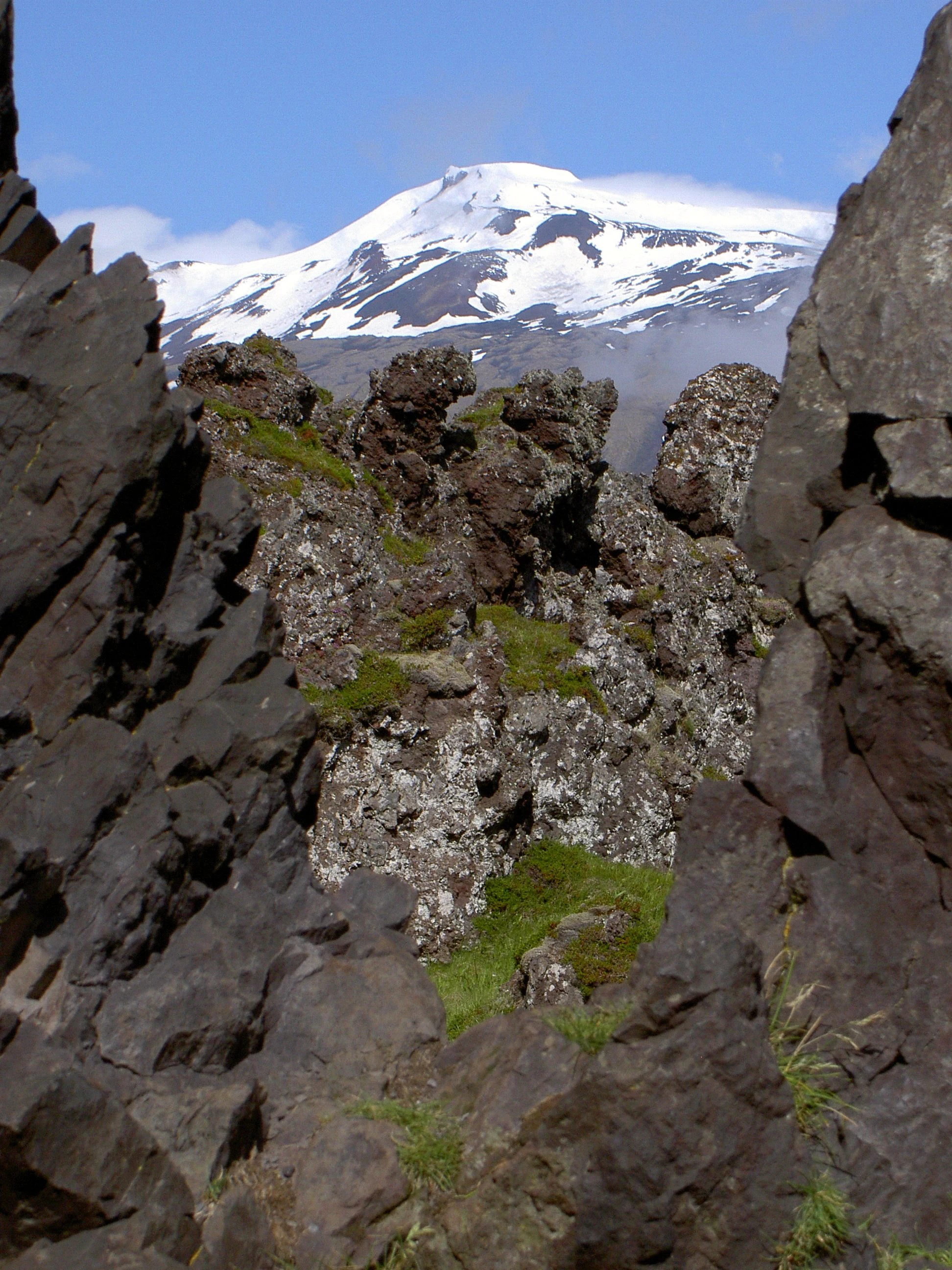 Una vista de un pico montañoso nevado y cubierto de glaciares, enmarcado por formaciones rocosas volcánicas oscuras y dentadas en primer plano.