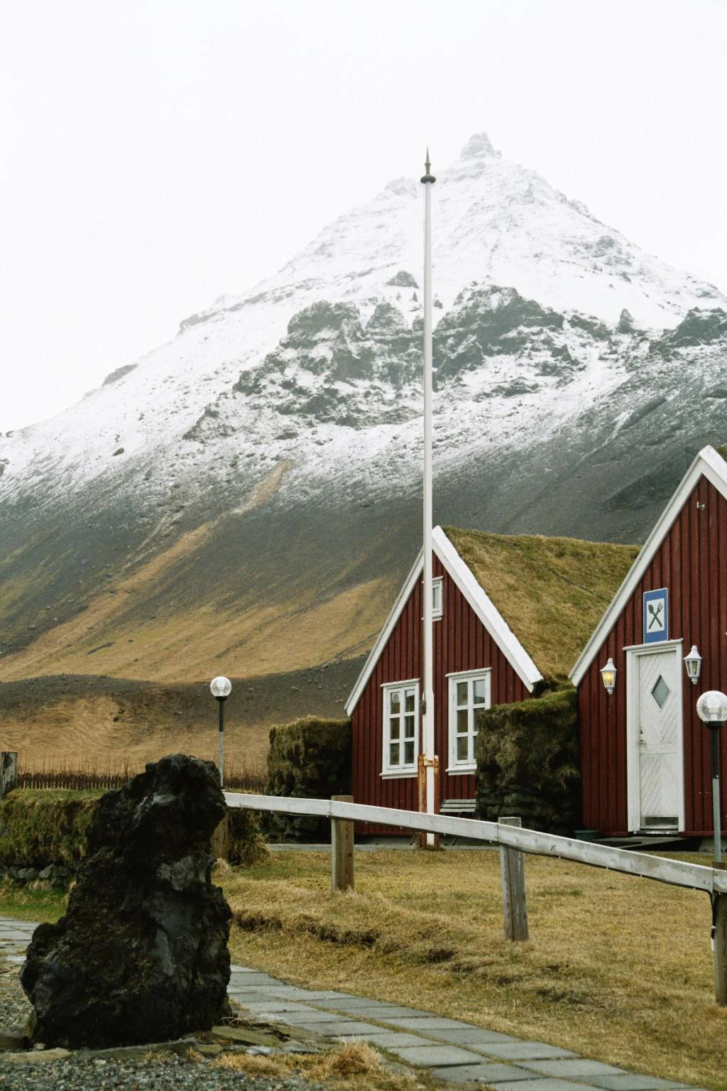 Two red wooden buildings with grass-covered roofs sit in the foreground, with a tall flagpole and a large volcanic rock; a snow-dusted mountain rises steeply in the background.