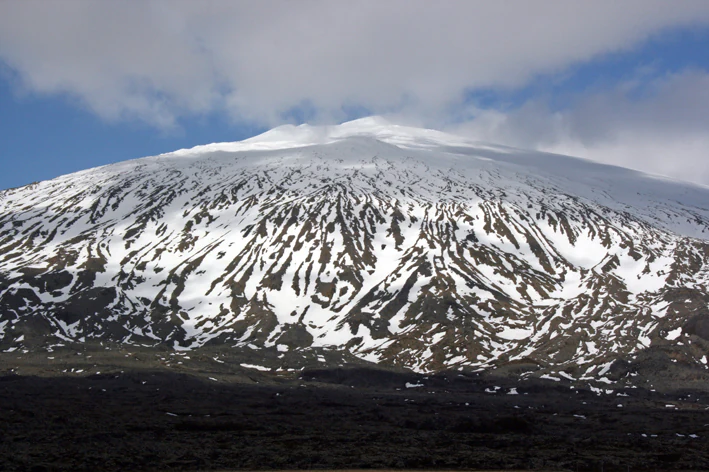 A wide view of the Snæfellsjökull volcano in Iceland, with snow covering its peak and upper slopes and dark volcanic rock at the base.