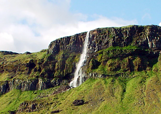 A thin waterfall flows down a steep, rocky green cliffside in Iceland.
