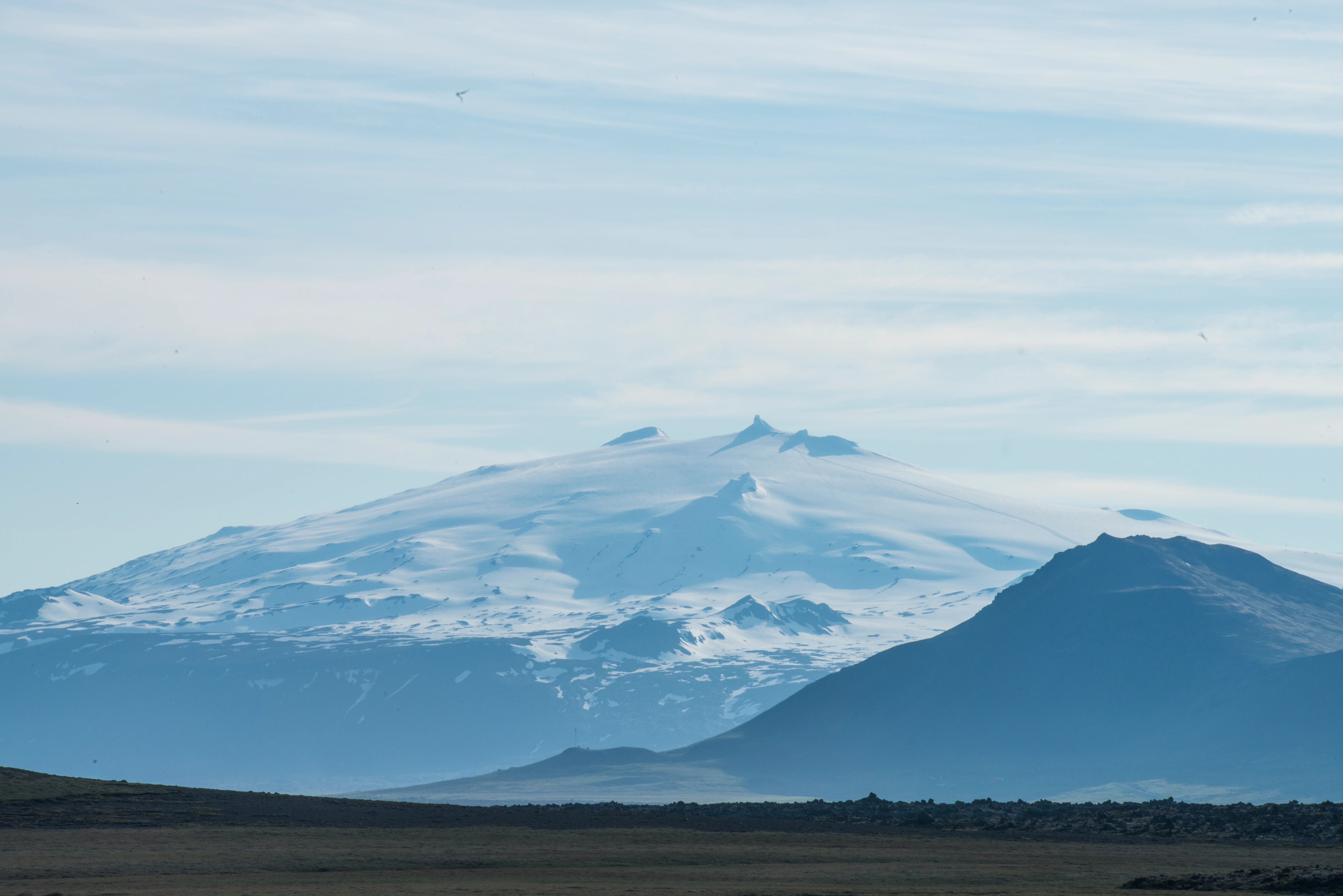 Snow-covered mountain peak with glacier, distant hills, and open landscape under a partly cloudy sky