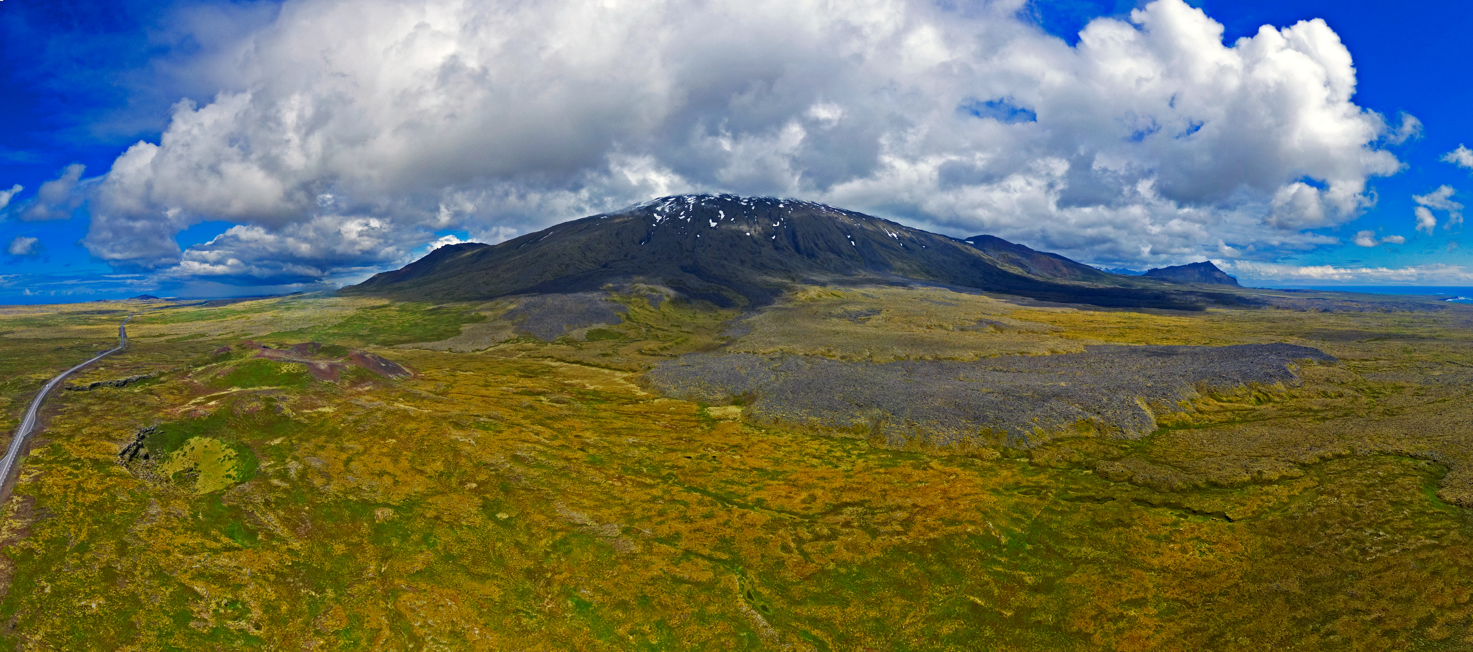Aerial view of Snæfellsjökull volcano showing snow-covered peak, rolling green and yellow fields, a winding road, and scattered clouds against a blue sky