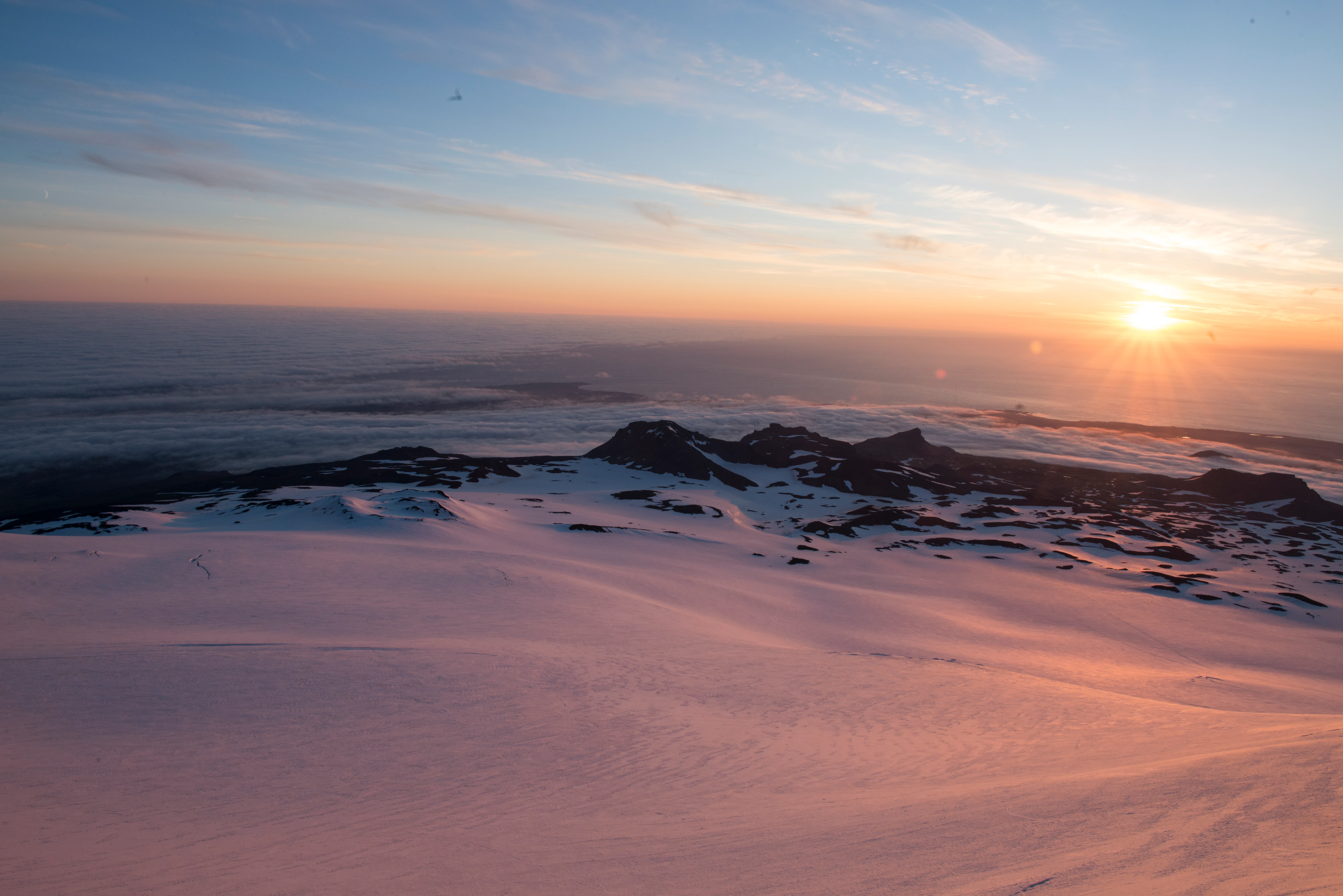 Snow-covered glacier landscape at sunset with the sun low on the horizon and distant sea visible