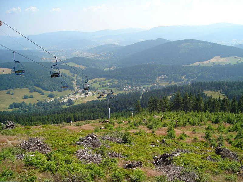 A high-angle view looking out over a mountain valley with a chairlift in the foreground, with rolling forested hills and mountain ranges in the distance under a hazy sky.
