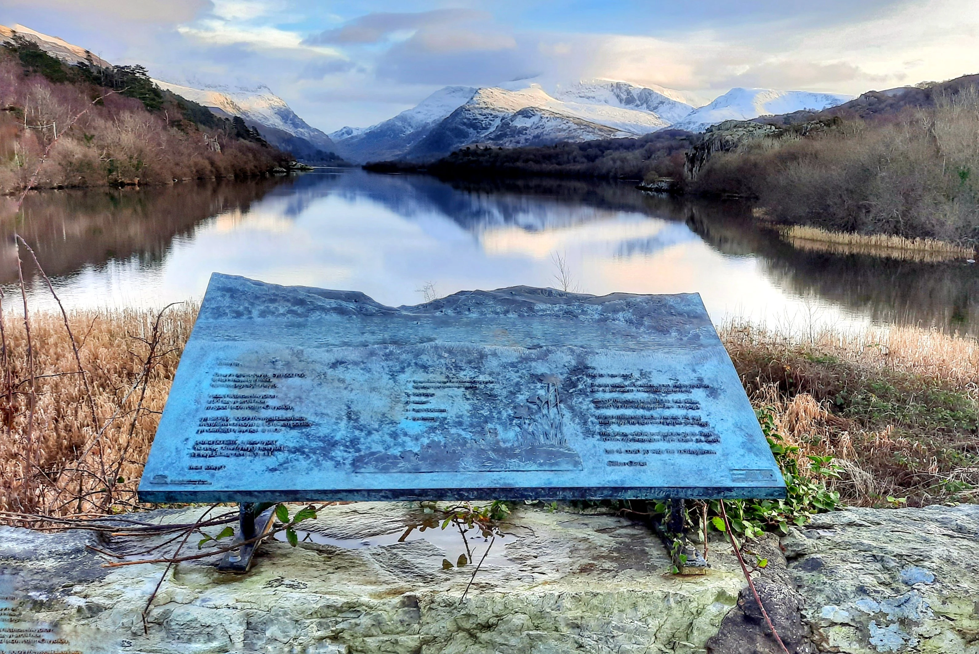 Blue informational sign with text and diagrams stands on a stone wall overlooking a calm lake reflecting mountains with snow-capped peaks