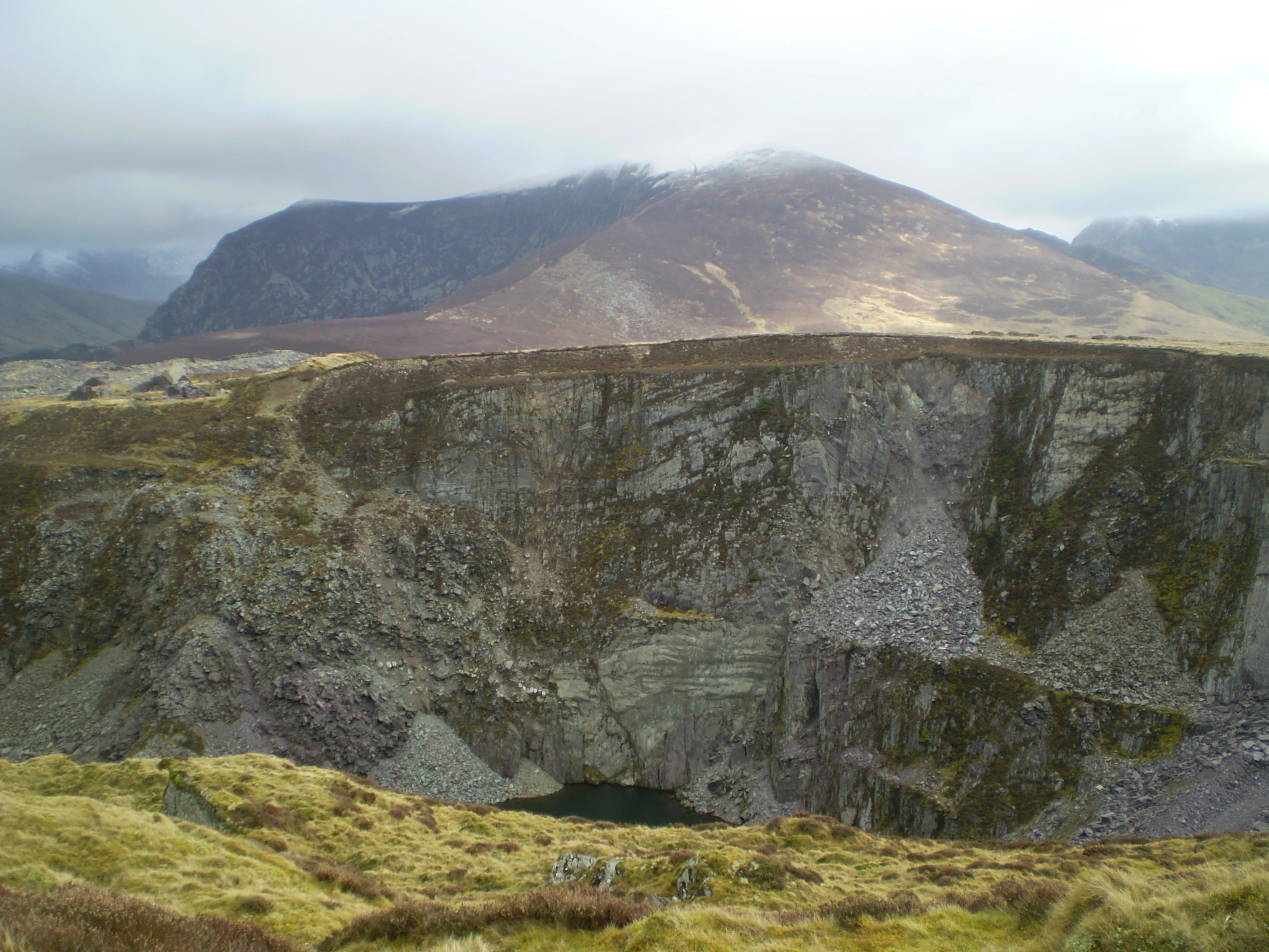 A wide view of a slate quarry with steep rocky cliffs and grassy terrain, with Mynydd Mawr mountain visible in the background under an overcast sky