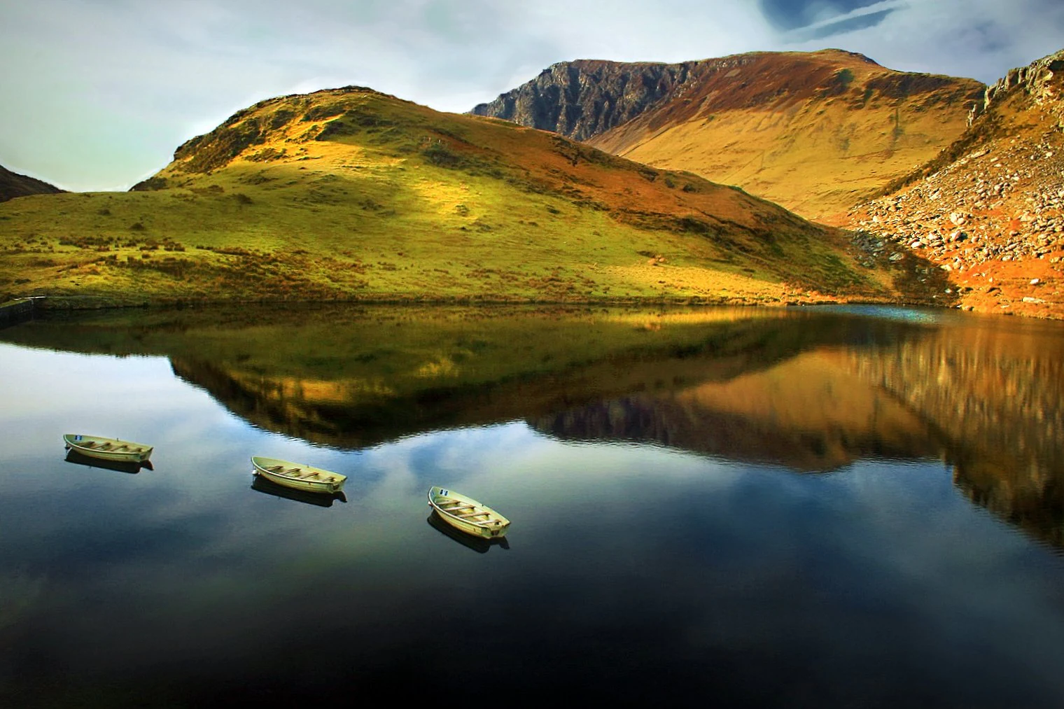 Three small boats floating on a still lake that perfectly reflects surrounding hills and mountains under a partly cloudy sky