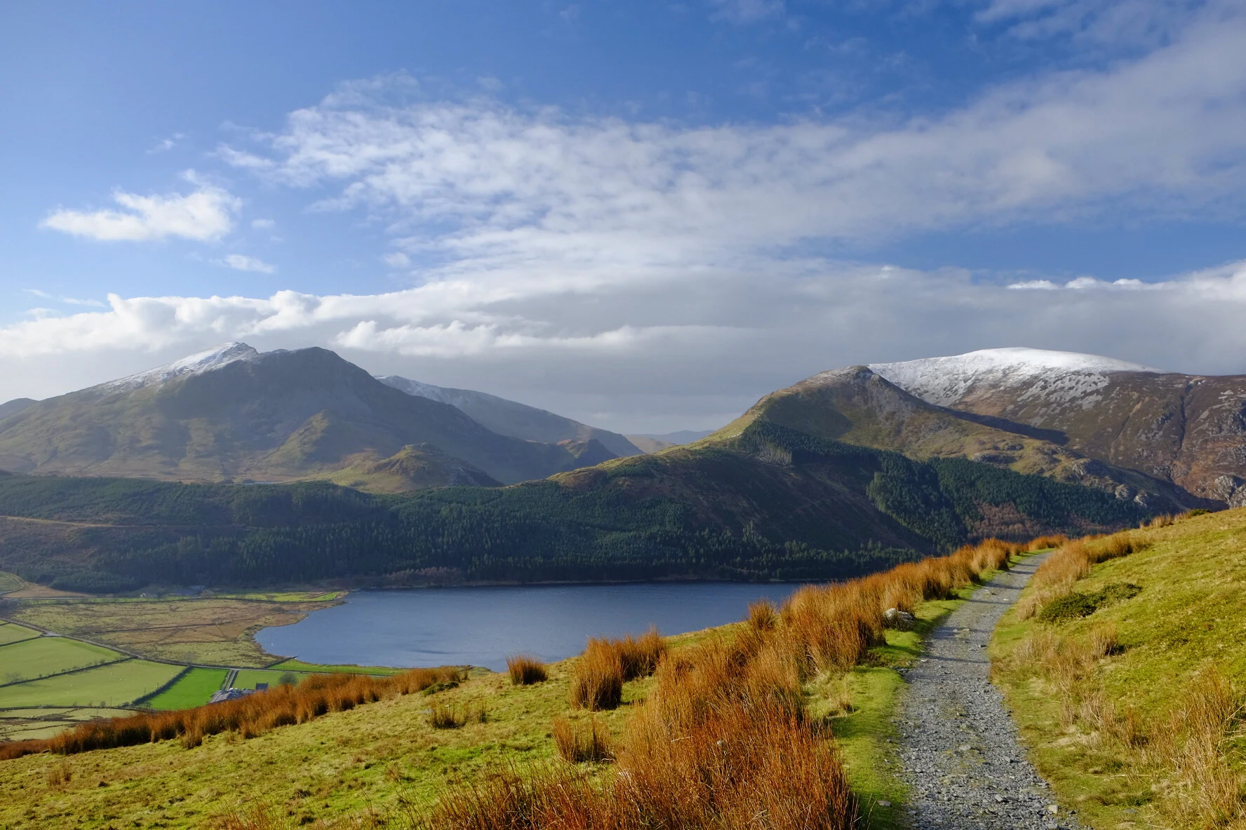 Gravel trail running through grassy hills with a lake and mountains in the background under a blue sky with clouds