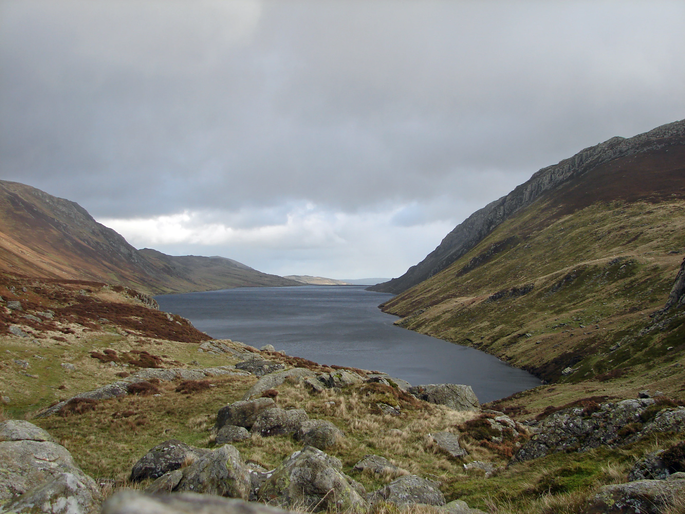 A lake surrounded by rocky hills and mountains under a cloudy sky with grassy and rocky terrain in the foreground.